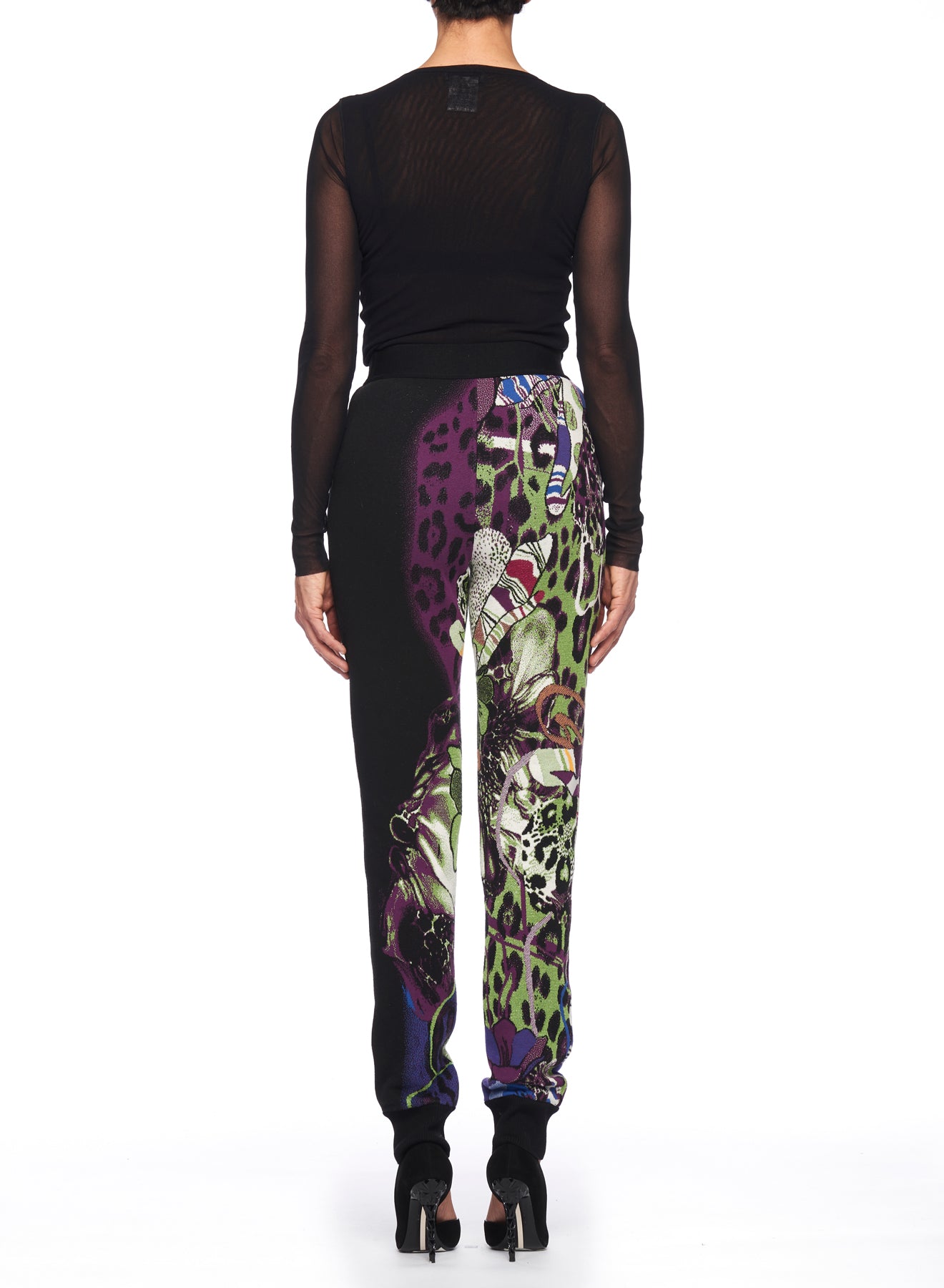 A person in a sheer black long-sleeve top and Fuzzi Woman Wild Side Joggers from Fuzzi, featuring a vintage-inspired colorful mixed print with purple and green accents, stands with their back to the camera. Their look is completed with black high-heeled shoes against a white background.