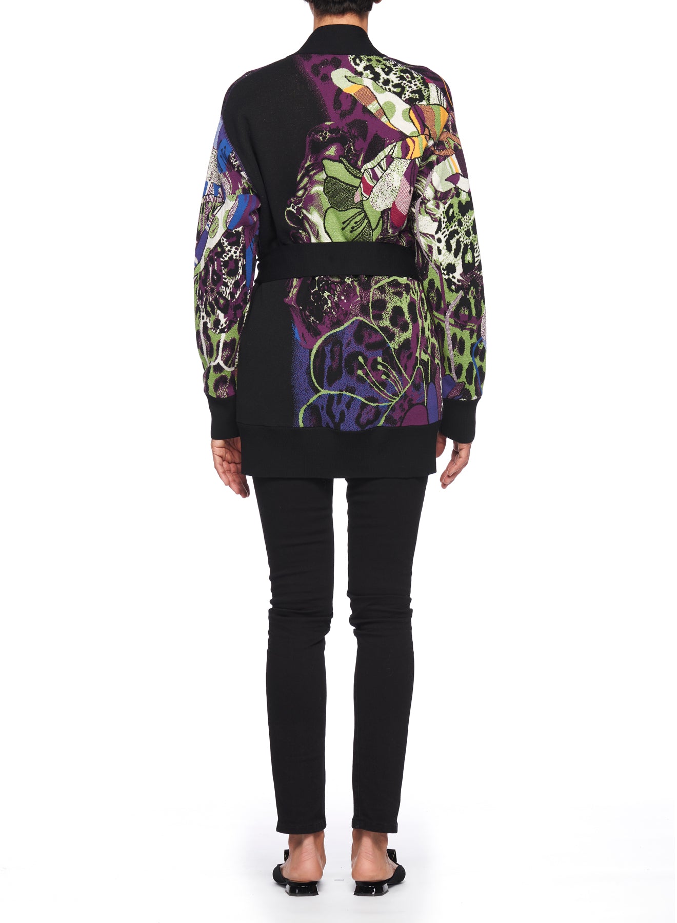 Wearing the Fuzzi Woman Eclectic Mix Cardigan adorned with vibrant insect and floral patterns, an individual stands facing away, complemented by black pants and sandals against a plain white backdrop.