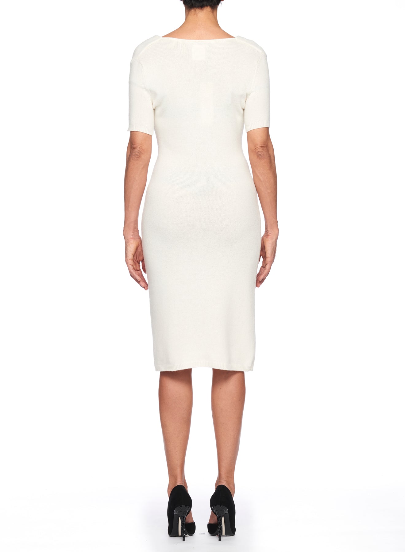 A person wearing the Fuzzi Woman White Cowl Neck Dress, a fitted, knee-length piece by Fuzzi, stands with their back to the camera. They are paired with black high-heeled shoes. The ribbed knit fabric enhances the texture against the plain white background.