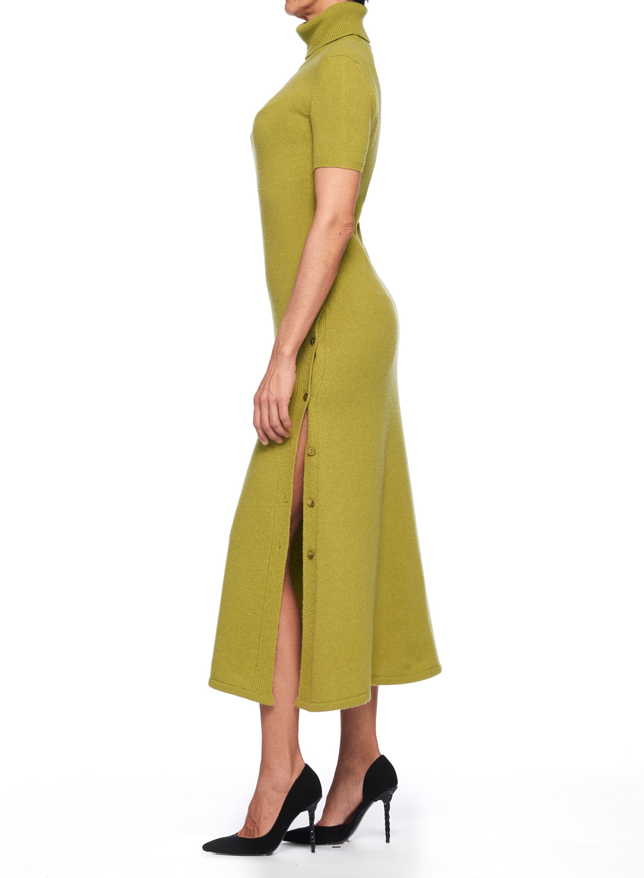 A person standing sideways in a Fuzzi Woman Dress with Side Slit, a light green, short-sleeved, ribbed knit sweater dress featuring button details on the side and a vintage look. The outfit is paired with black high heels against a plain white background.