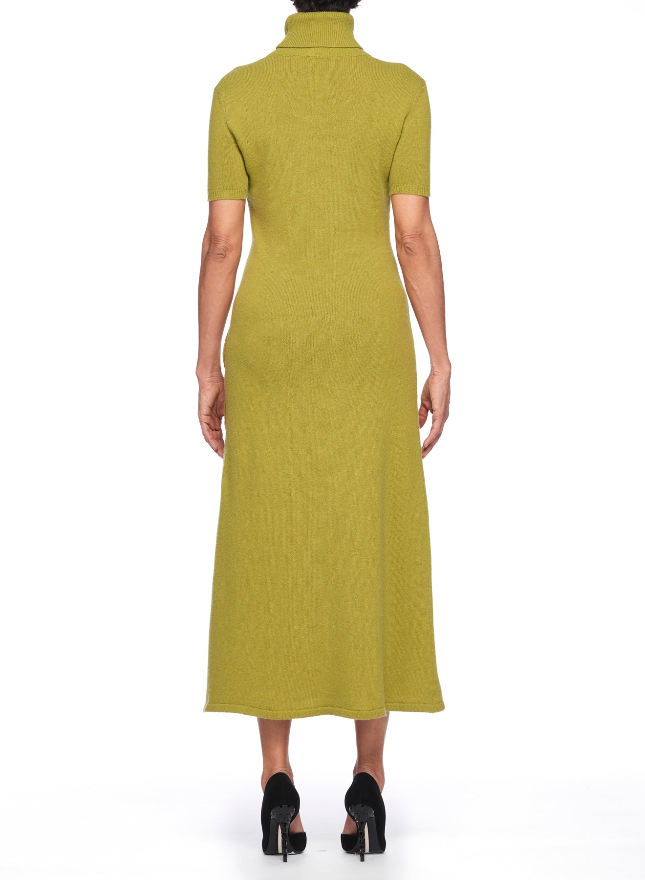 A person models a Fuzzi Woman Dress with Side Slit, characterized by its vintage olive green hue and short-sleeved turtleneck design, crafted from ribbed knit fabric. They stylishly pair the dress with black heels against a plain white backdrop.