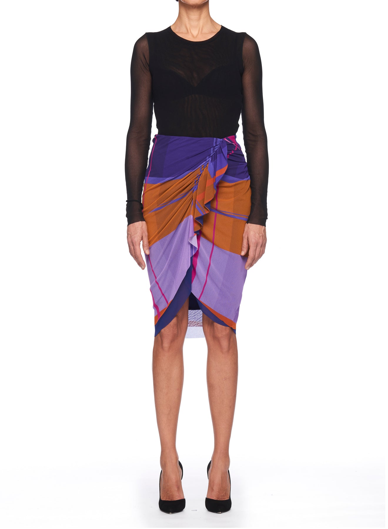 A person stands against a white background wearing a black sheer long-sleeve top, paired with the Fuzzi Woman Colorful Wrap Skirt with Ruffle Detail, showcasing vibrant orange, purple, and blue geometric patterns. They complete the look with black heels.