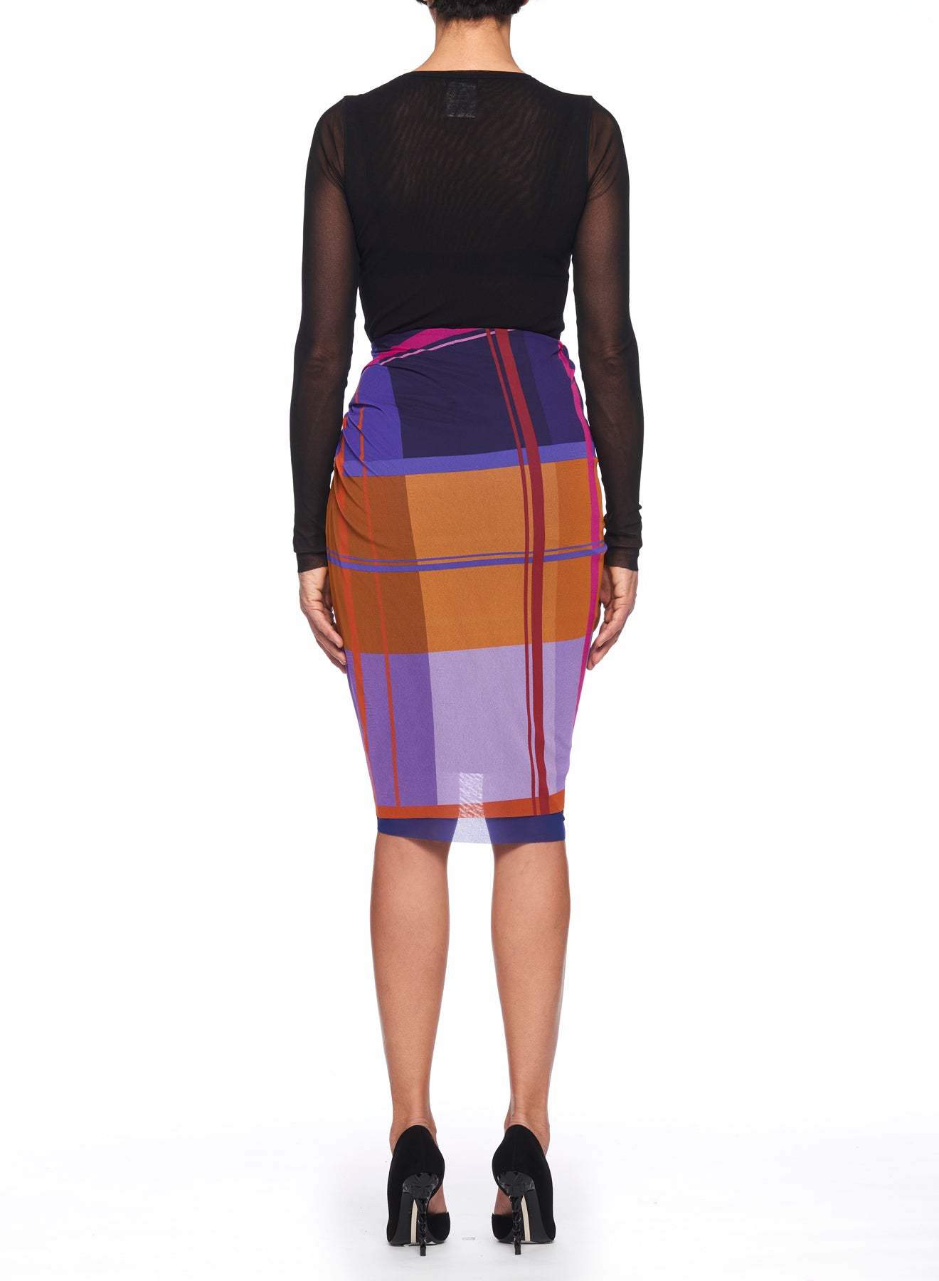 A person in a sheer black long-sleeve top and a Fuzzi Woman Colorful Wrap Skirt with Ruffle Detail, serving as a vintage statement piece, stands facing away. They are wearing black high-heeled shoes against a white background.