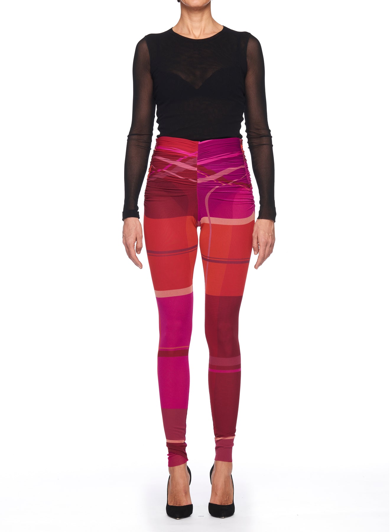 Wearing a black sheer long-sleeve top, an individual poses against a white background, showcasing the Fuzzi Woman Abstract Print Leggings by Fuzzi. These vibrant leggings feature high-waisted geometric patterns in shades of red and pink, elegantly paired with stylish black high heels.