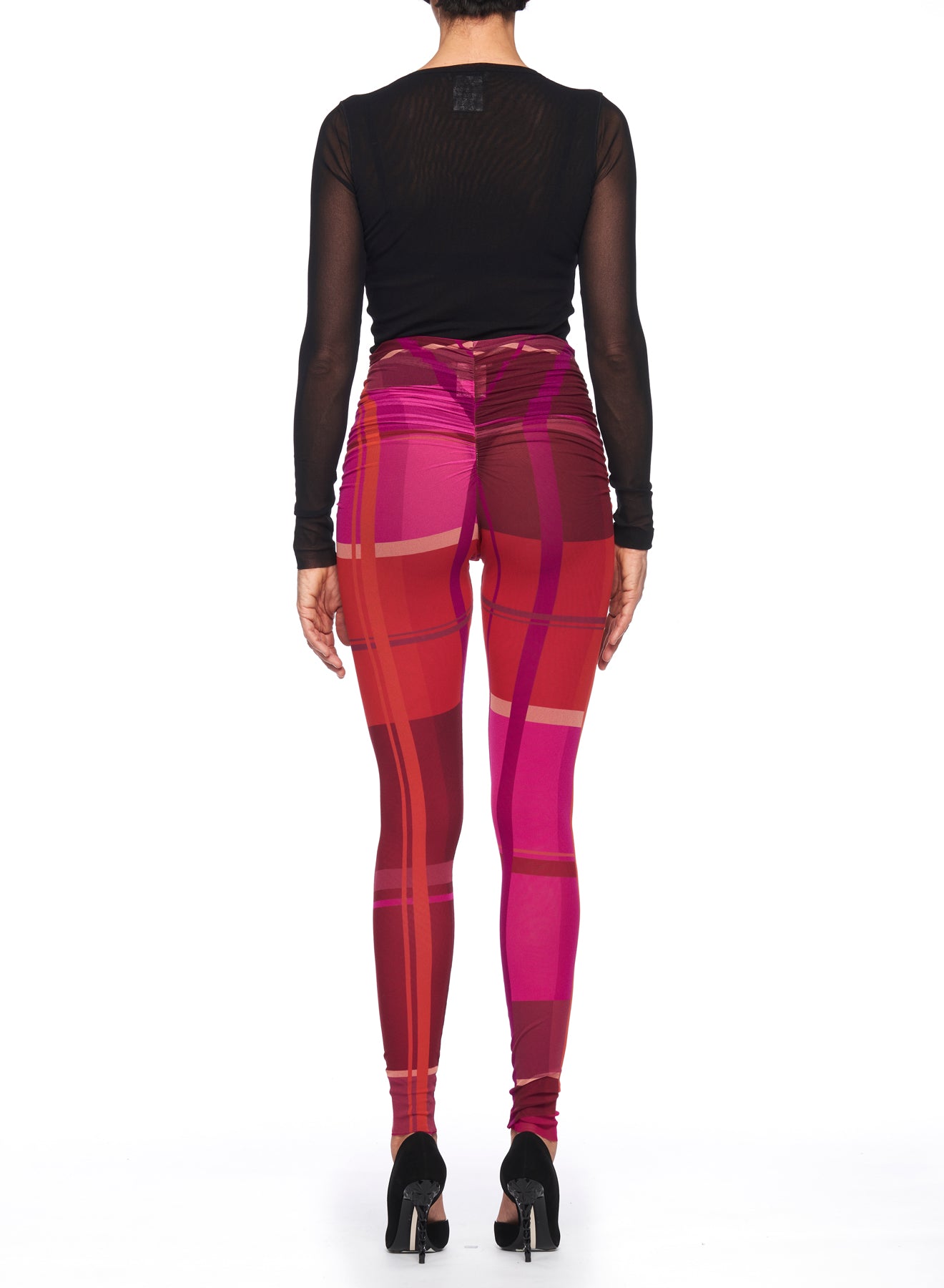 A person facing away is styled in a sheer black long-sleeve top, paired with high-waisted, vibrant Fuzzi Woman Abstract Print Leggings from the brand Fuzzi, featuring shades of pink and red plaid. Their look is completed with sleek black high heels.