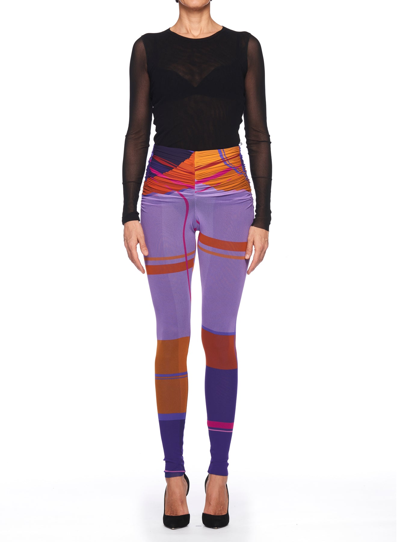 A person stands against a white background wearing a black sheer long-sleeve top, sleek black high heels, and vibrant Fuzzi Woman Abstract Print Leggings by Fuzzi, which feature an eye-catching high-waisted fit with orange, purple, and red patterns.