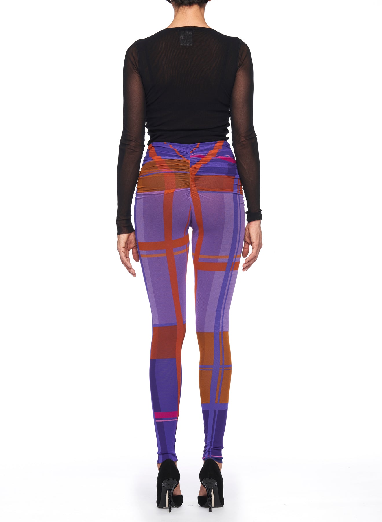 A person wearing a long-sleeved black mesh top and Fuzzi Woman Abstract Print Leggings with an abstract design in purple, orange, and pink tones stands with their back to the camera. They are also wearing black high-heeled shoes.