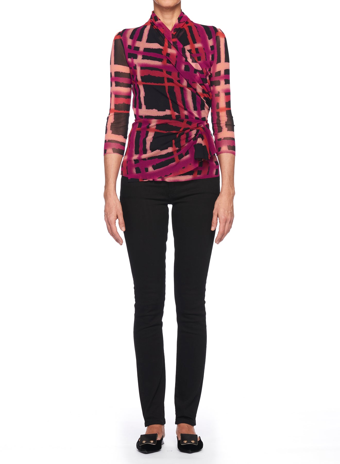 A person stands against a white background wearing the Fuzzi Woman Abstract Print Top with Tie Waist by Fuzzi, which features a sheer, vintage aesthetic and an abstract design in shades of pink, purple, and black. The top showcases a wrap-around style and three-quarter length sleeves. They have paired it with black pants and black flat shoes.