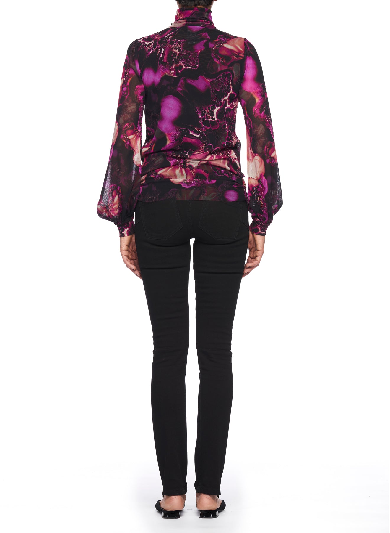 A person wearing the Fuzzi Woman Blouse with Puff Sleeves and Ruched Detailing, featuring a sheer abstract print in purple and pink hues, is paired with black skinny pants and shoes, standing with their back to the camera on a white background. Their hair is elegantly tied up.