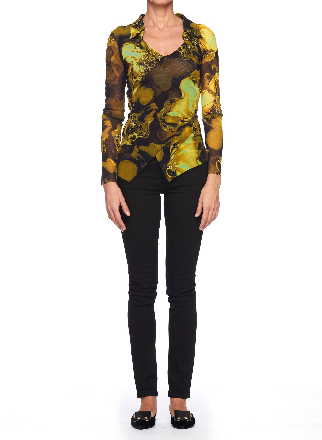 A person stands against a white background wearing the Fuzzi Woman Print Mesh Top with V-Neck, featuring a long-sleeve multicolored abstract pattern in shades of yellow and brown, paired with black pants and shoes. The image is cropped at the neck.