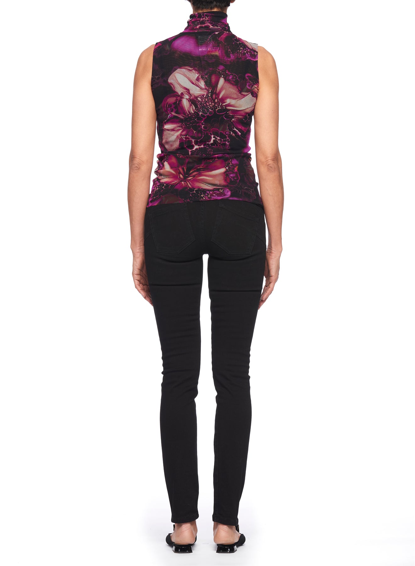 A person is standing with their back to the camera, wearing a Fuzzi Woman Top with Sleeveless Design featuring a vintage floral pattern of pink and purple flowers, paired with black jeans. Their arms are relaxed by their sides against a plain white background.