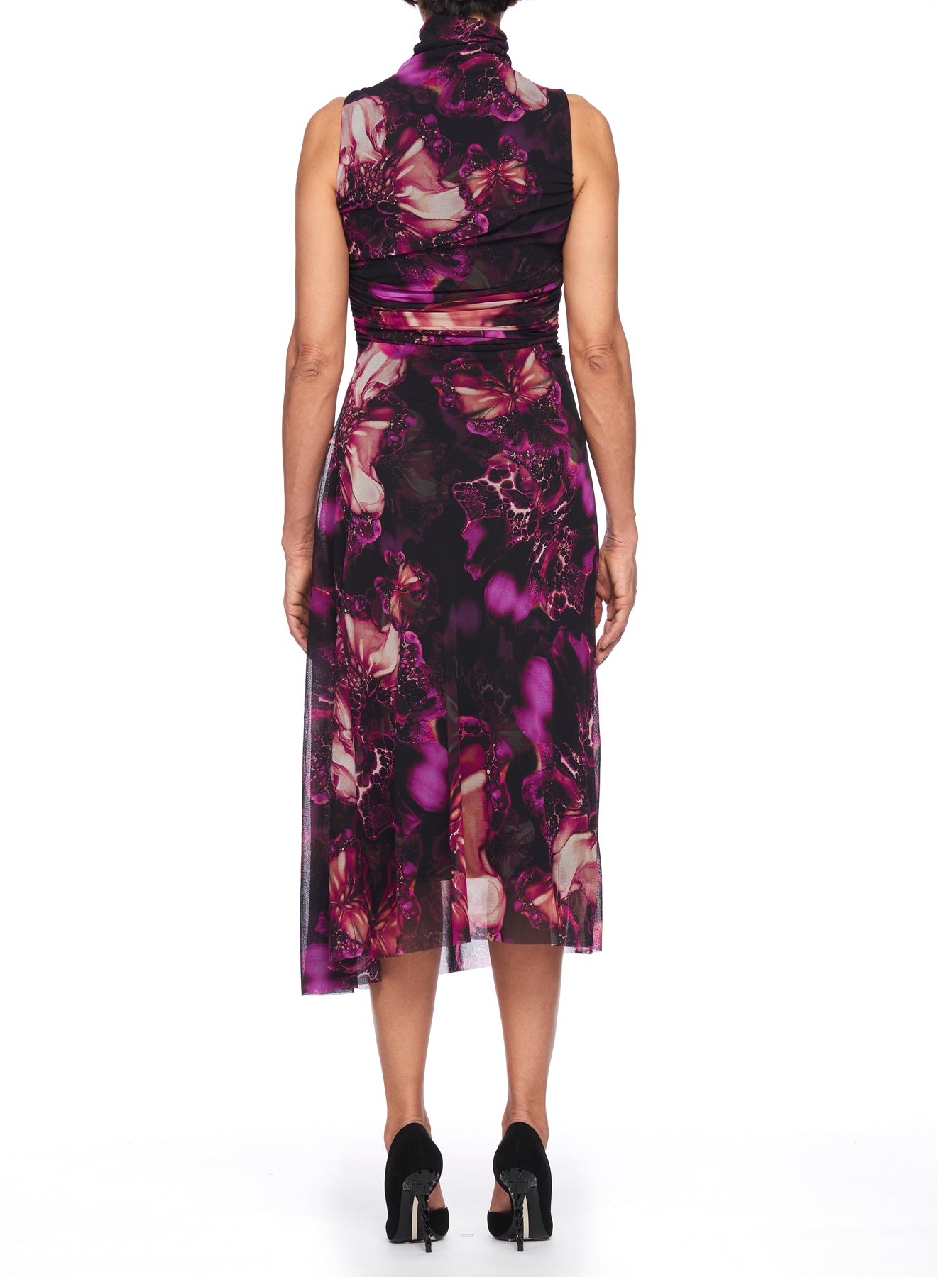 A woman is wearing the Fuzzi Woman Mesh Dress with High Neck, a knee-length piece featuring a purple floral pattern reminiscent of vintage designs. She stands facing away, paired with black high-heeled shoes against a white background.