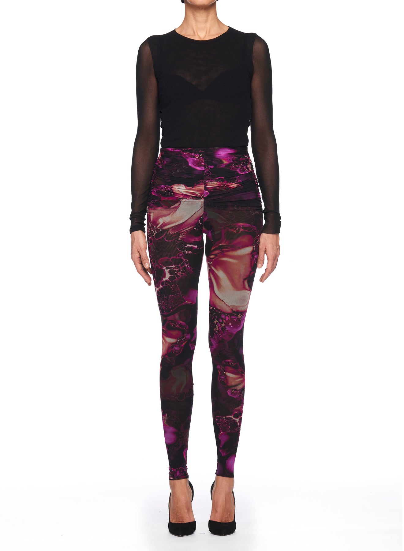 A person is modeling their ensemble against a white background, wearing a black long-sleeve top with sheer sleeves and Fuzzi Woman Leggings with Intricate Pattern from the brand Fuzzi, accentuated by vibrant purple and pink abstract designs. The outfit is completed with black high-heeled shoes.