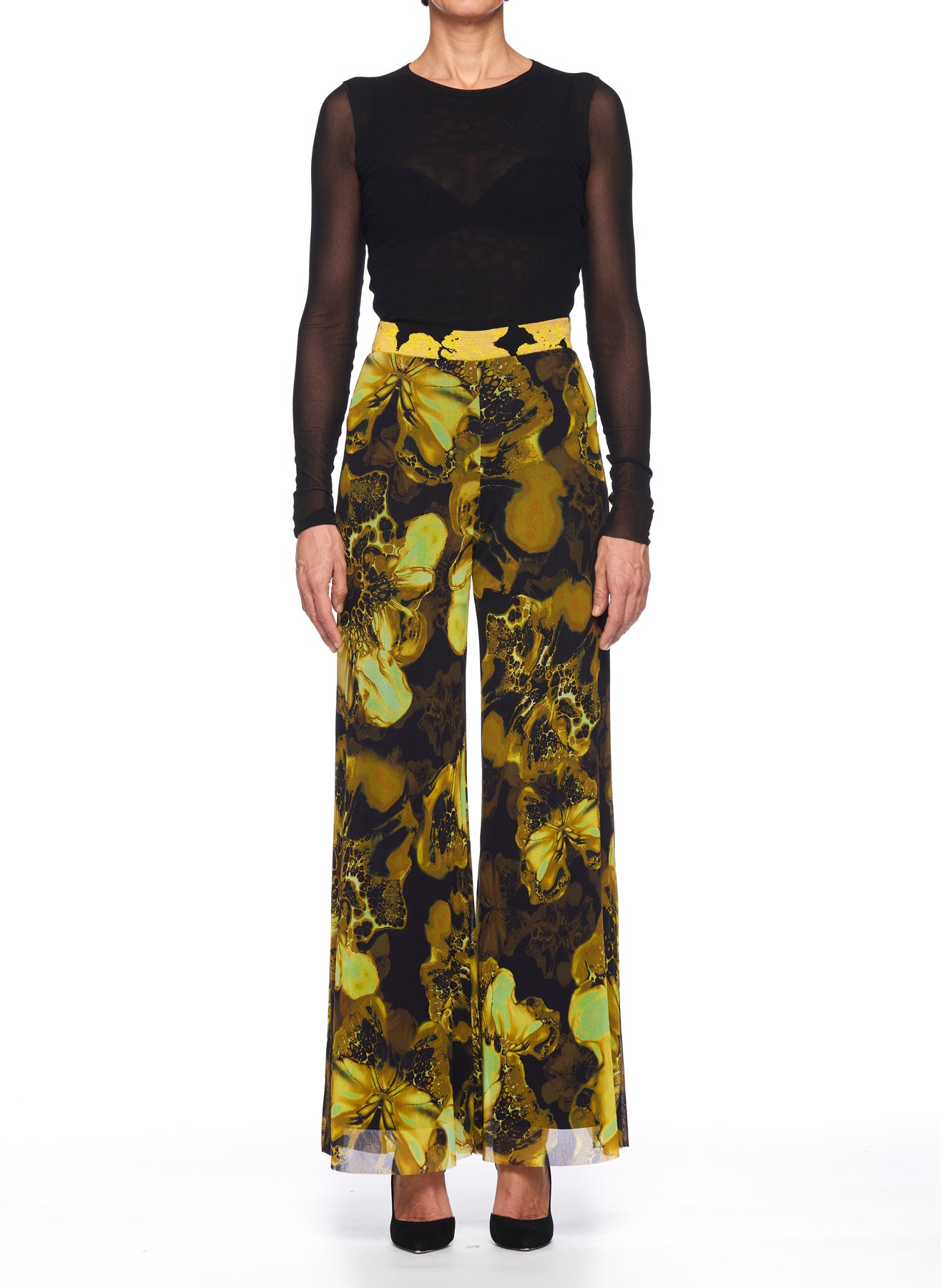 A person dressed in a sheer black long-sleeve top and the Fuzzi Woman Statement Pants, featuring a high-waisted design and an abstract yellow and black floral pattern, stands against a white background with visible black heels.