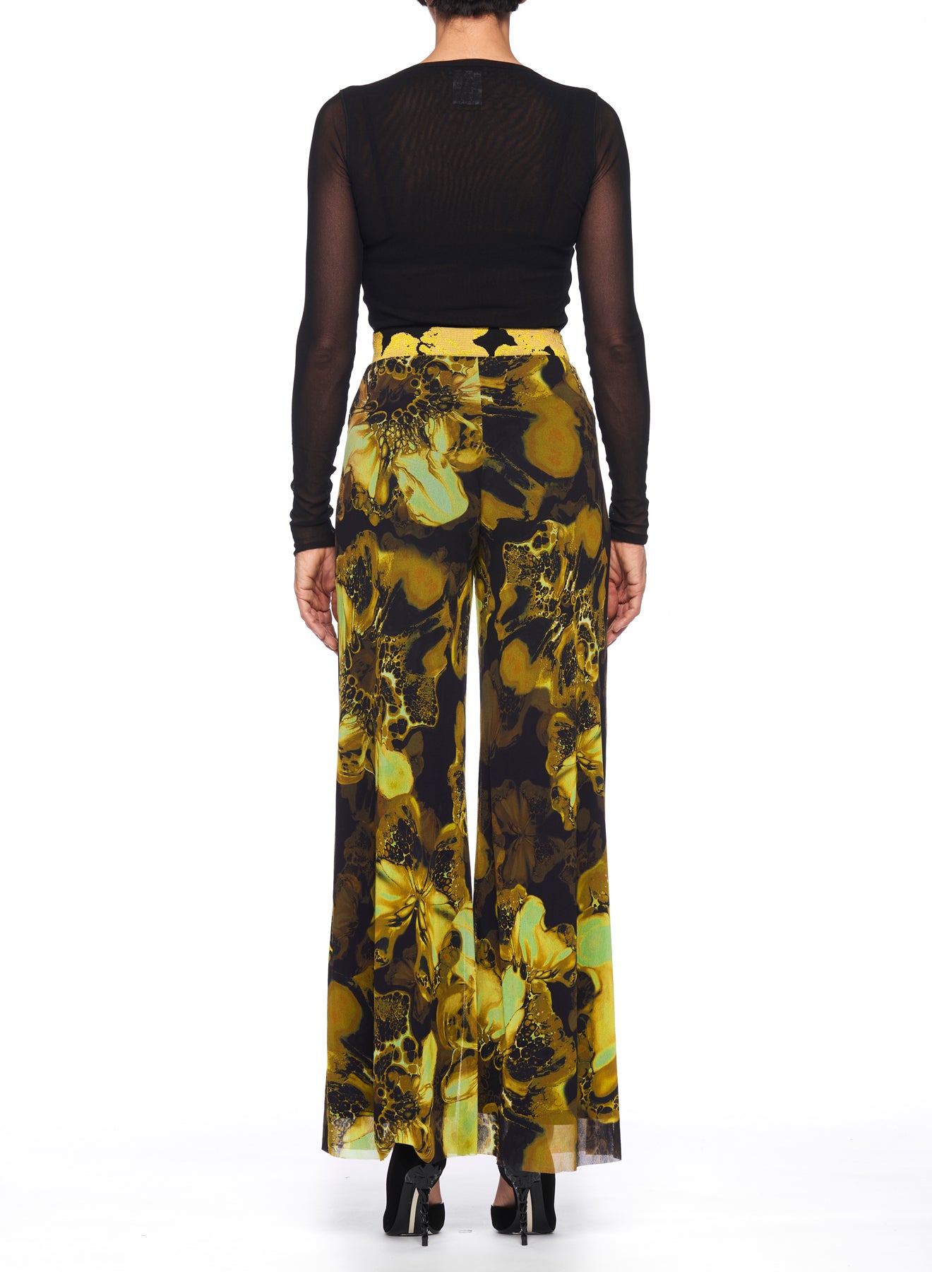 A person stands facing away, highlighting the Fuzzi Woman Statement Pants with High-Waisted Design, featuring an abstract yellow and black floral print. The look is completed with a black sheer long-sleeve top and black heels against a plain white backdrop.