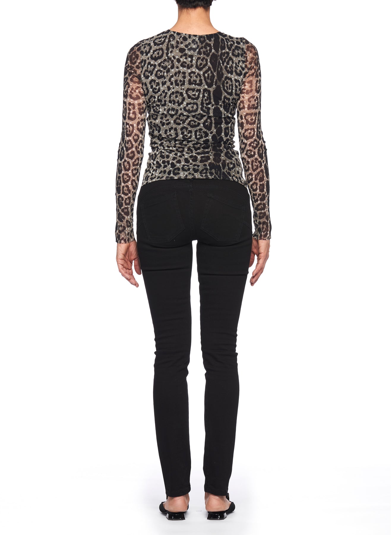 Rear view of a person standing against a white backdrop. They are wearing the Fuzzi Woman Leopard Print Top with Long Sleeves, crafted from sheer fabric, paired with black skinny jeans and sandals. Their arms rest naturally by their sides.