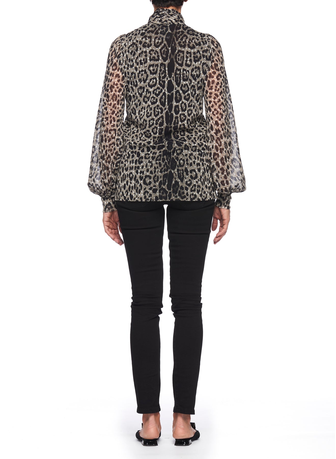 A woman sporting the Fuzzi Woman Edgy Mesh Top, which features a chic leopard print and sheer long sleeves, pairs it with black fitted pants and black shoes while standing facing away. The backdrop is a simple white canvas.