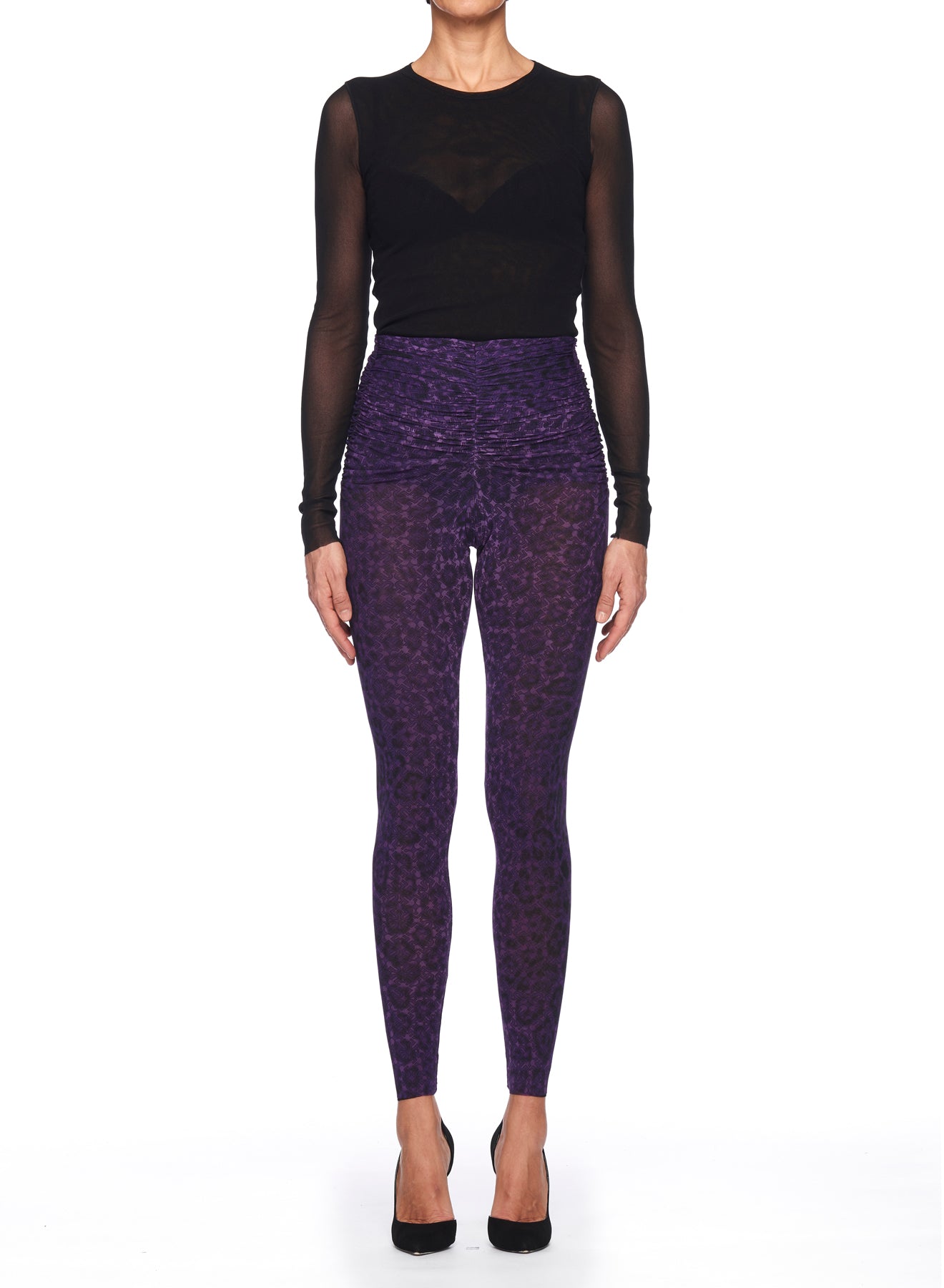 A woman stands against a white background, wearing a black sheer long-sleeve top and the Fuzzi Woman Leopard Print Pants from Fuzzi in vibrant purple. She completes her vintage-inspired look with black high heels, her arms relaxed by her sides.