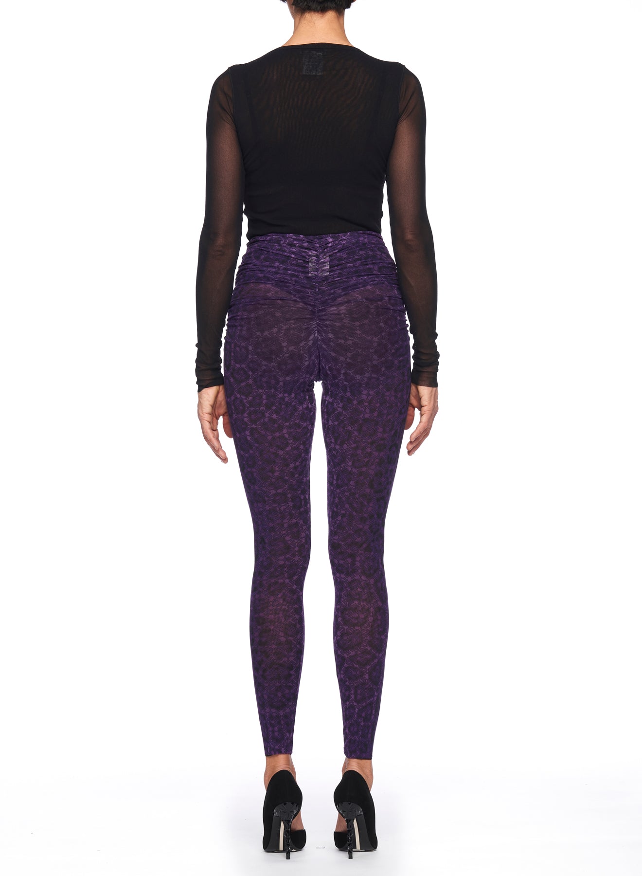 A person wearing the Fuzzi Woman Leopard Print Pants is seen from the rear, showcasing striking purple leopard print leggings. The high-waisted design by Fuzzi accentuates their silhouette and is paired with elegant black high-heeled shoes, all set against a plain white background that highlights this chic ensemble.