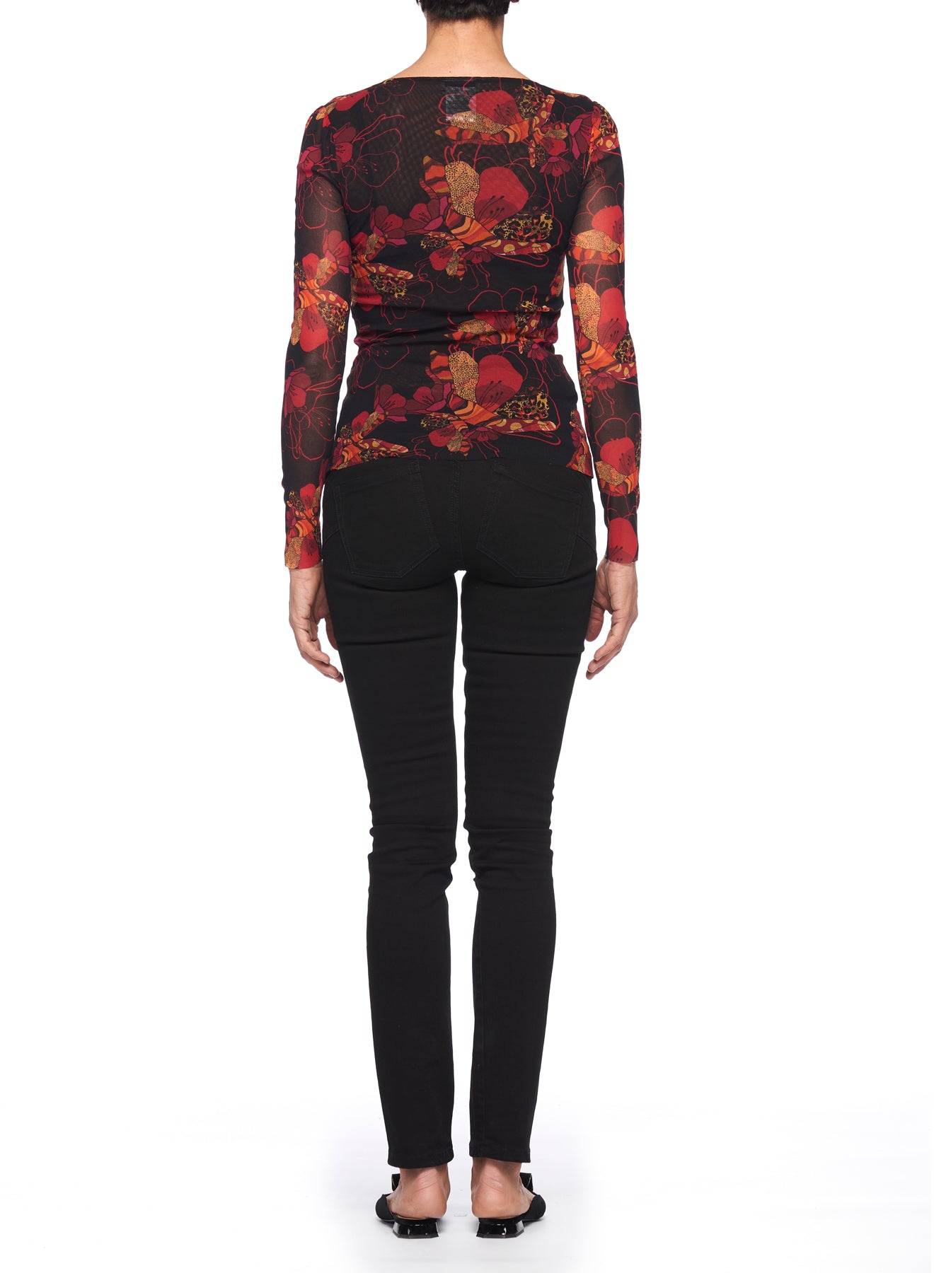 Rear view of a person standing against a white background, wearing the Fuzzi Woman Mesh Top by Fuzzi, featuring sheer fabric with floral patterns in red and orange tones, paired with black jeans and black flats.