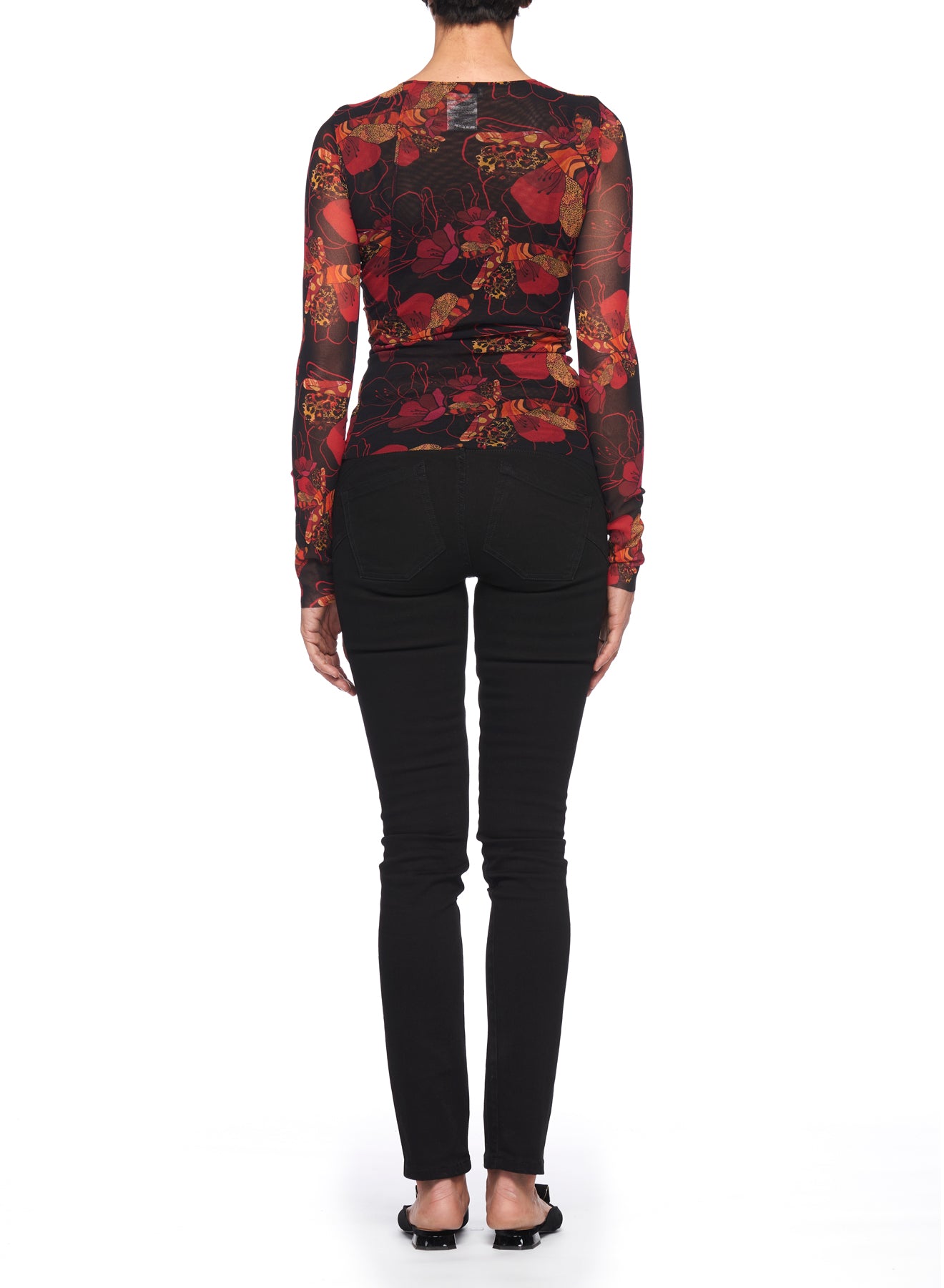 The individual stands with their back to the camera, highlighting the Fuzzi Woman Floral Print Mesh Top flawlessly paired with black jeans. Their ensemble is completed by black flat shoes set against a plain white background.