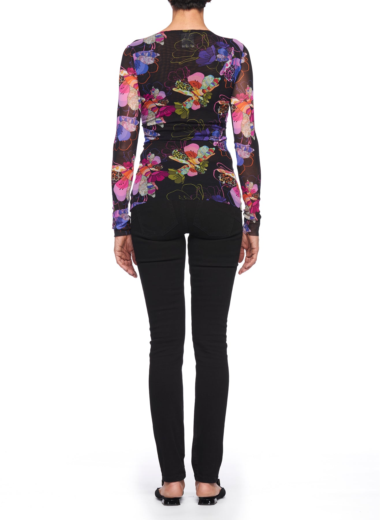 The individual faces away from the camera, showcasing the Fuzzi Woman Boho-Chic Floral Mesh Top, featuring a vibrant floral pattern. They complement their outfit with black pants and shoes, all set against a simple white background.