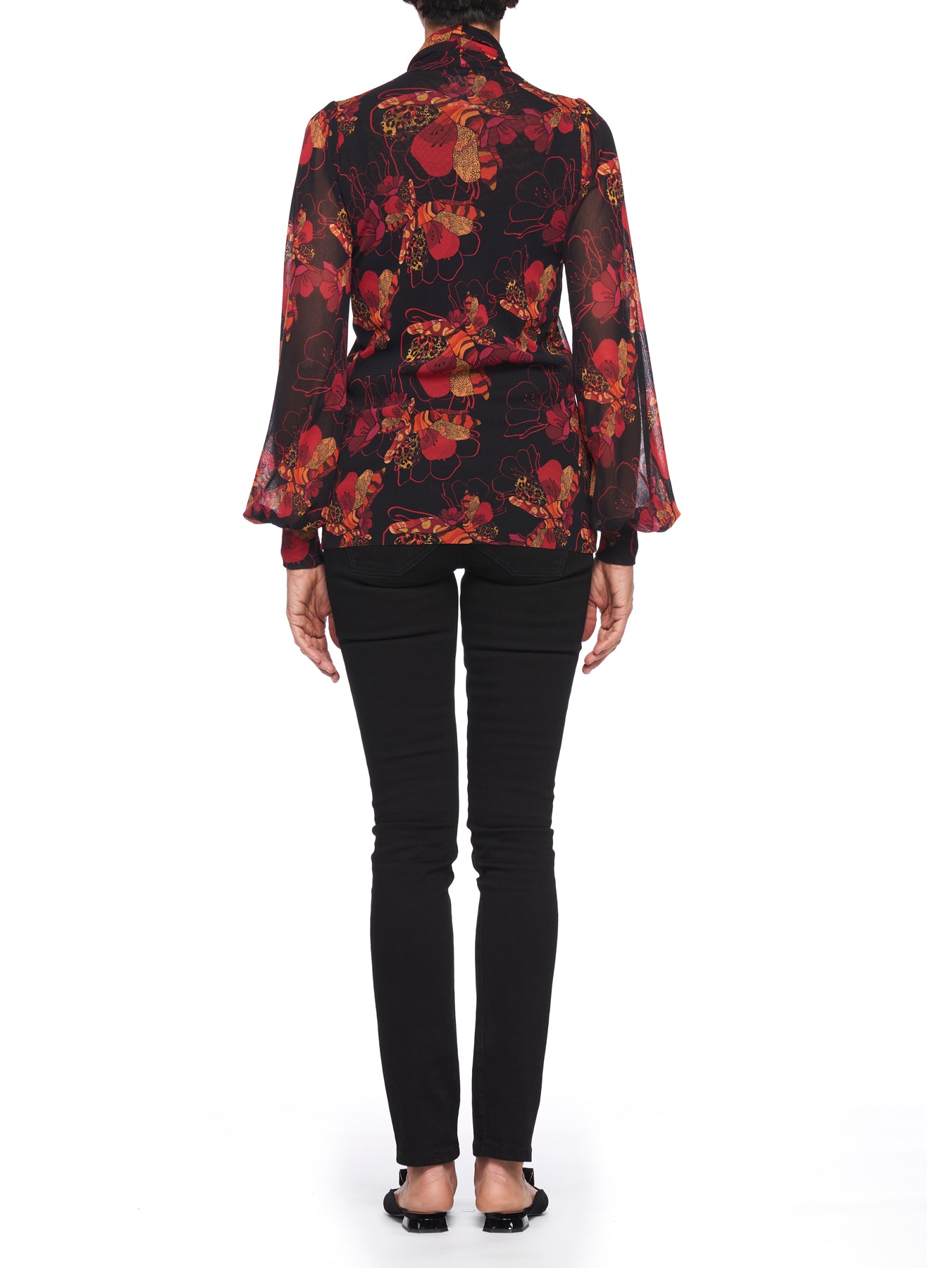 Rear view of a person wearing the Fuzzi Woman Romantic Floral Print Blouse with Tie Neck and black pants. The blouse features sheer fabric adorned with vintage-style red and orange designs on a black background. The individual stands against a white backdrop, wearing black shoes.