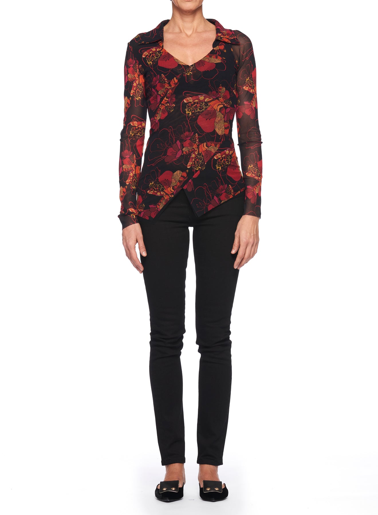 A person stands against a plain white background, wearing the Fuzzi Woman V-neckline Top with an abstract print in dark red and black, paired with black pants and ballet flats. Made by Fuzzi from sheer mesh fabric, the outfit exudes elegance as they face forward with arms relaxed by their sides.