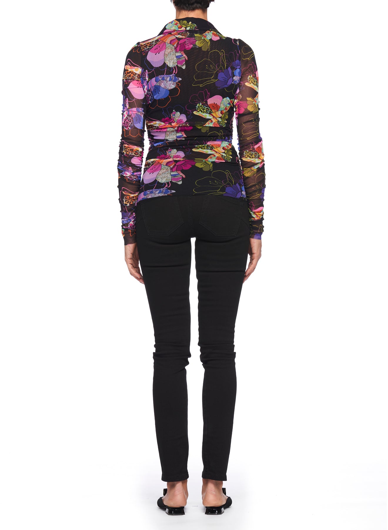 A person stands facing away, wearing the Fuzzi Woman Boho-Chic Mesh Top by Fuzzi, featuring a vibrant floral print and paired with black fitted pants. They are sporting black open-toed sandals against a plain white background.