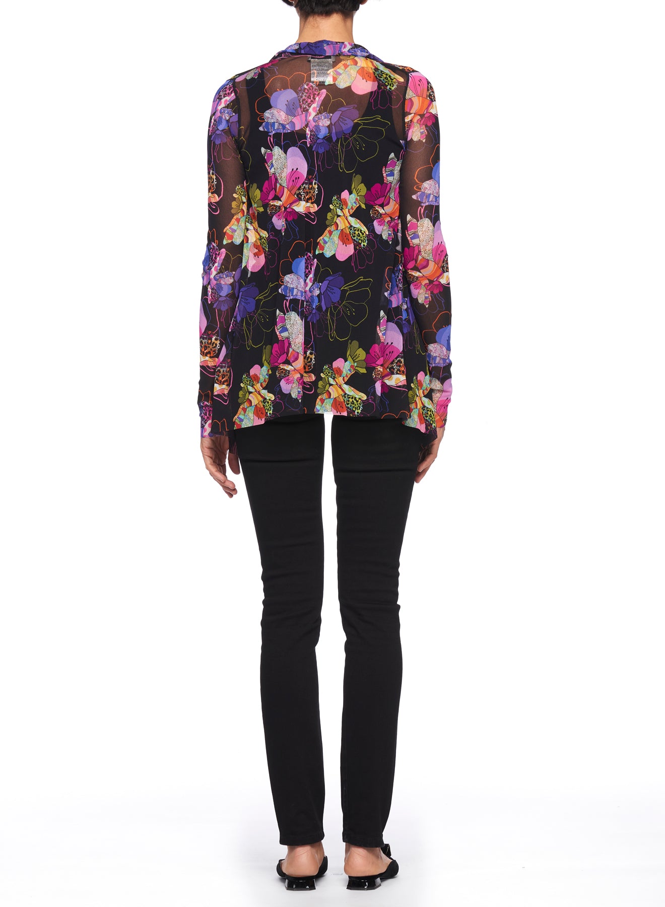 A person is seen from the back wearing the Fuzzi Woman Boho-Chic Floral Mesh Cardigan, which features a sheer, long-sleeve design adorned with a vibrant floral print on a black background. They have paired it elegantly with black pants and shoes, all set against a simple white backdrop.