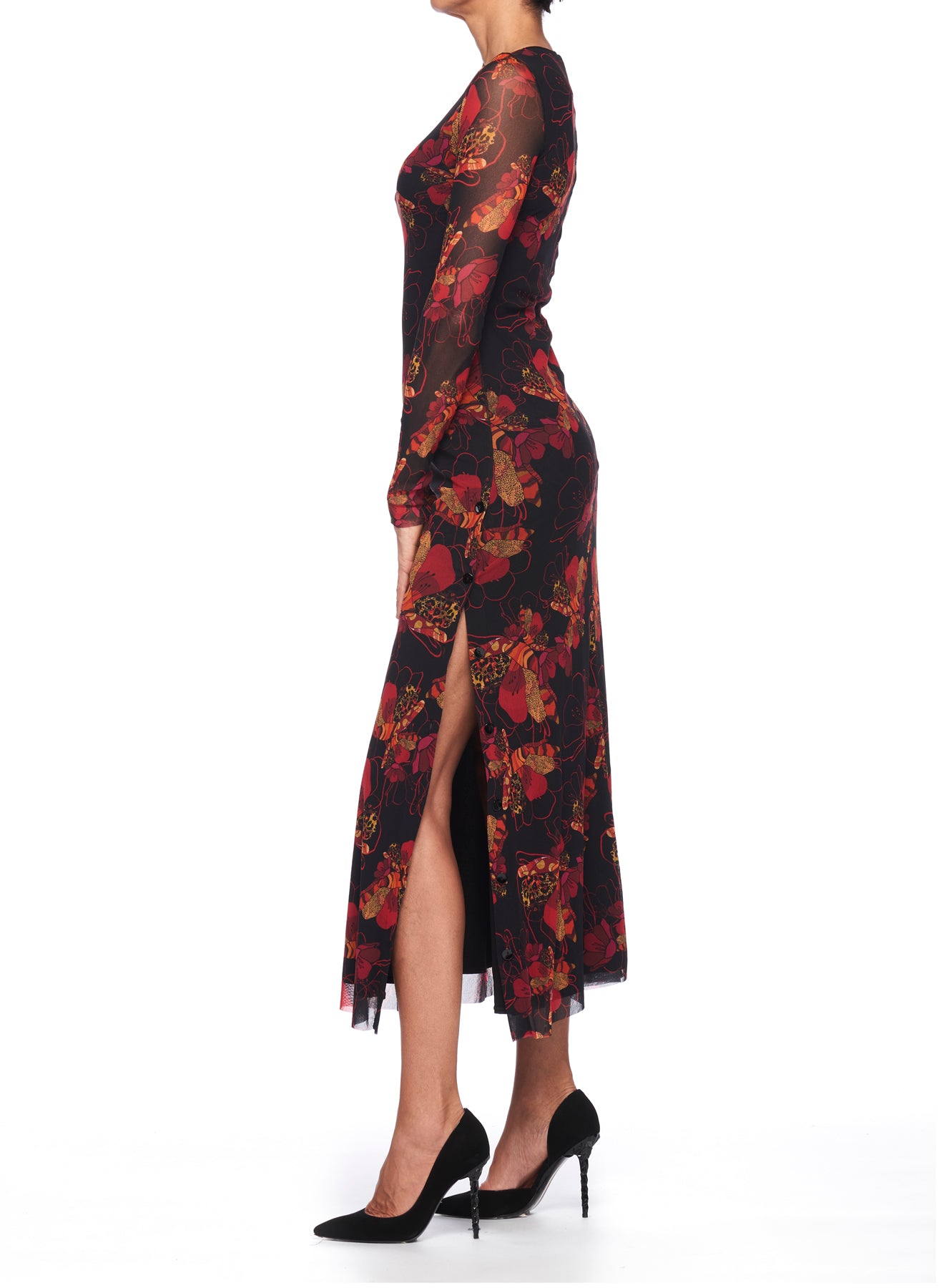 A person is shown in profile against a white background wearing the Fuzzi Woman Romantic Floral Mesh Dress, which showcases vibrant red and orange flowers in a vintage floral print. The midi dress is styled with a side slit and is perfectly paired with black high heels for an elegant look.