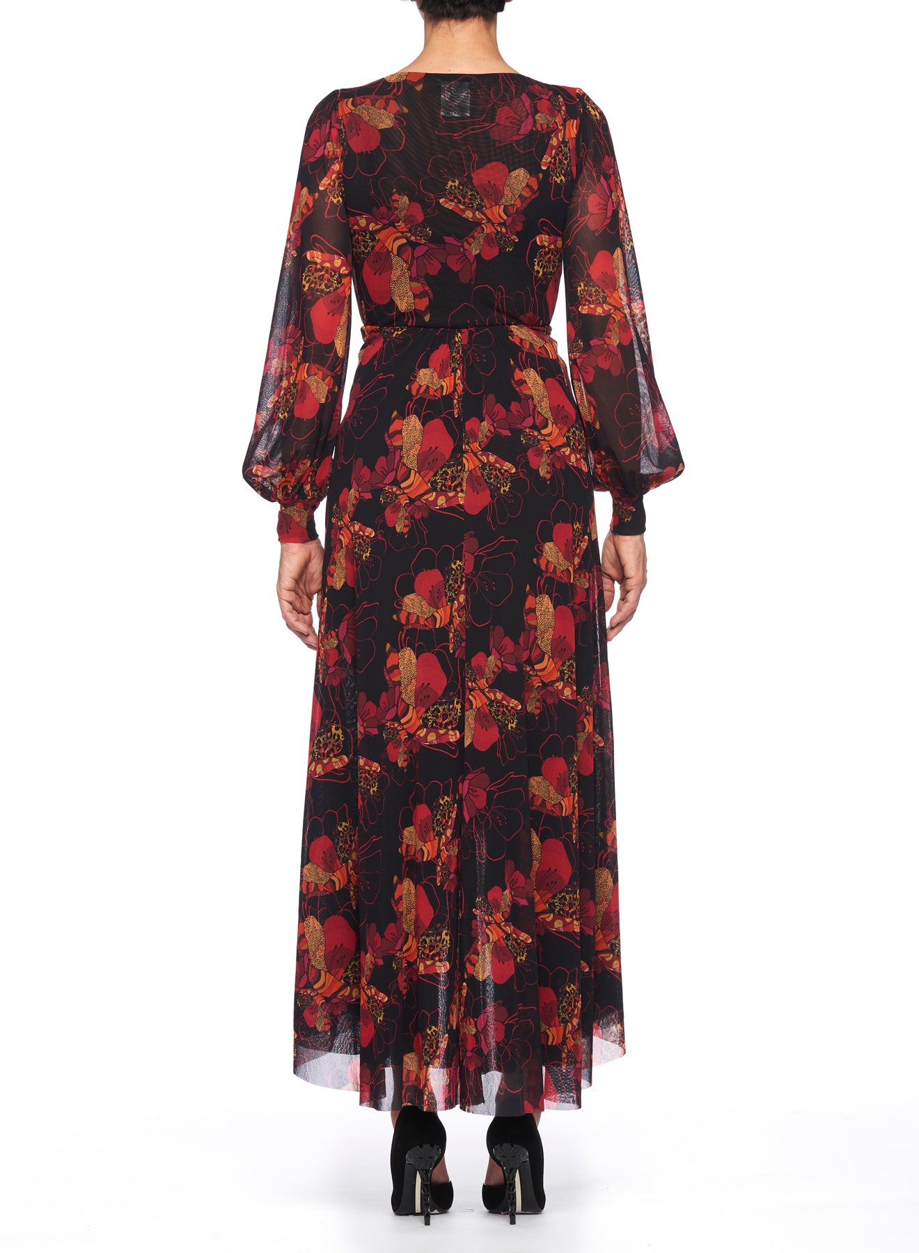 A person is seen from behind wearing the Fuzzi Woman Dramatic Floral Wrap Dress, a long, sheer black dress adorned with red floral patterns. This sophisticated wrap silhouette is complemented by long, flowy sleeves. They have paired the outfit with black heels.