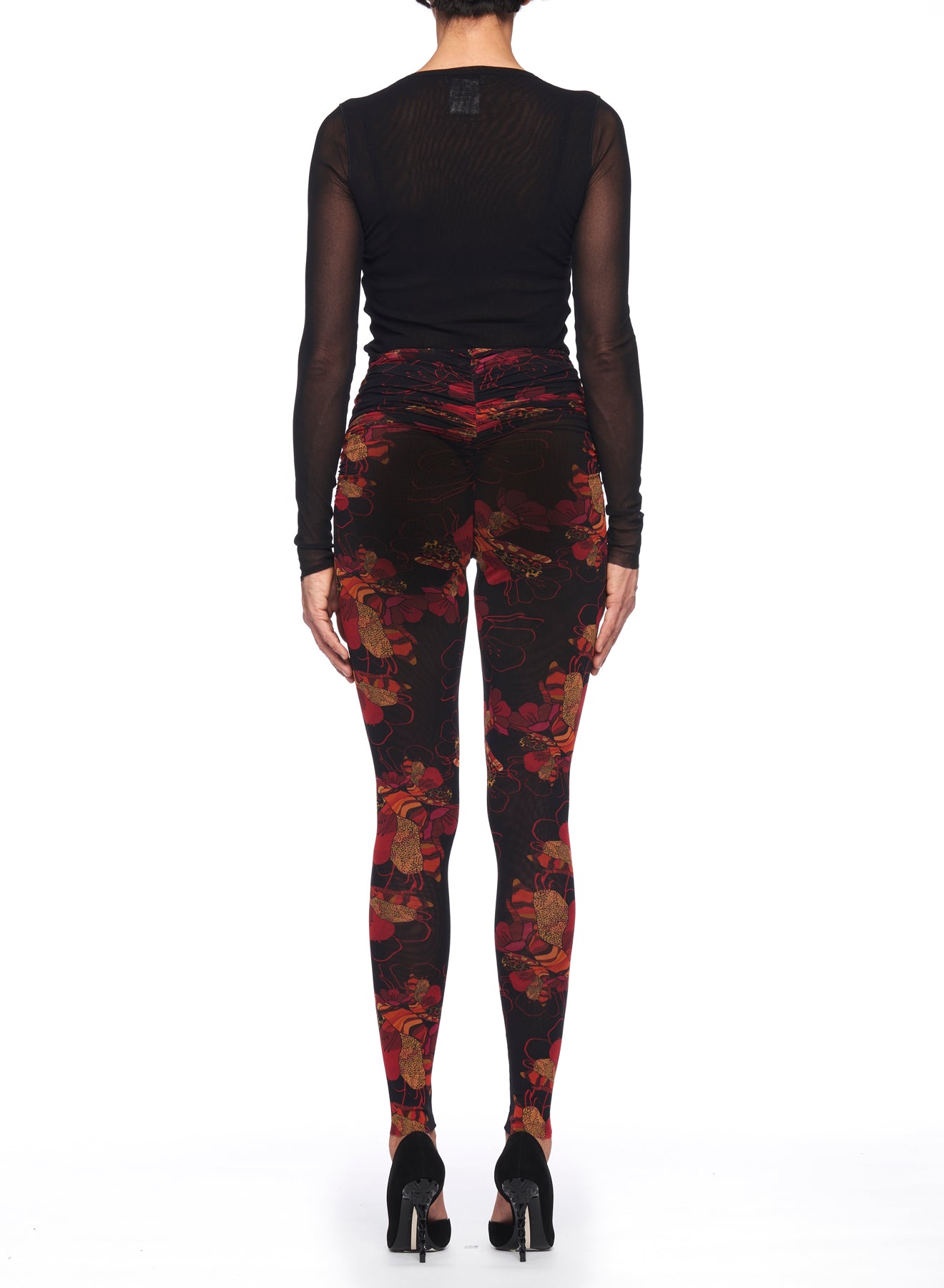 A person stands with their back to the camera, wearing a black mesh long-sleeve top and high heels. The vibrant floral design of the Fuzzi Woman Floral Leggings contrasts beautifully against the plain white background.