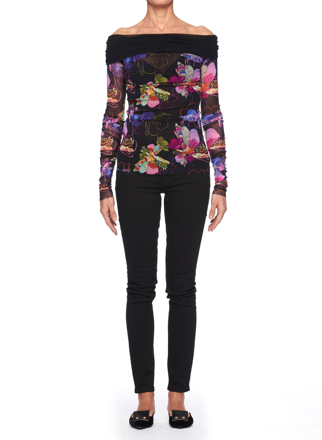 A person wearing the Fuzzi Woman Off-Shoulder Floral Mesh Top, featuring sheer mesh fabric and a colorful floral print, is paired with black pants and shoes, standing against a plain white background.