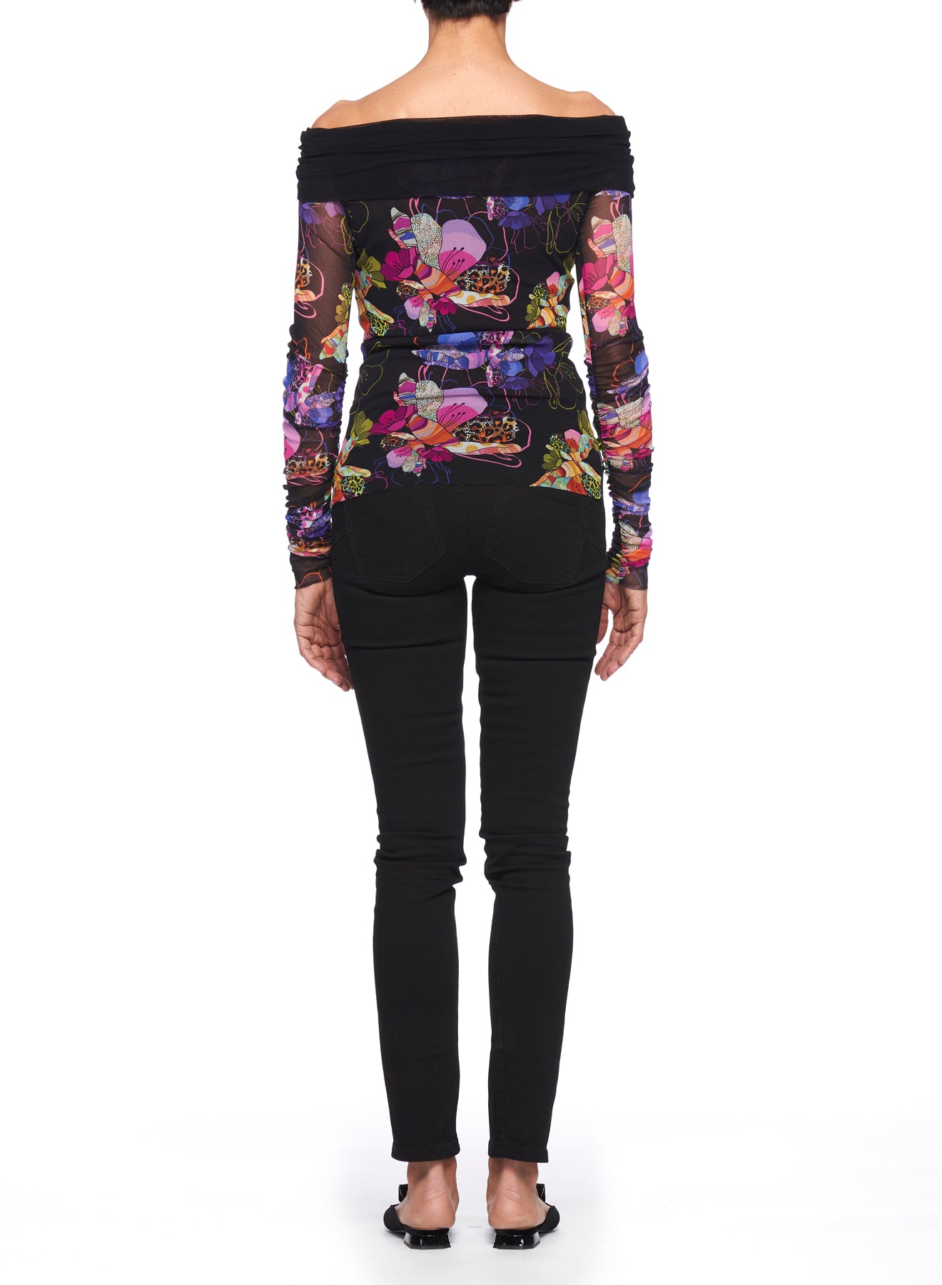 Rear view of a person in the Fuzzi Woman Off-Shoulder Floral Mesh Top, featuring vibrant floral prints and sheer mesh fabric, paired with black pants, standing against a white background.