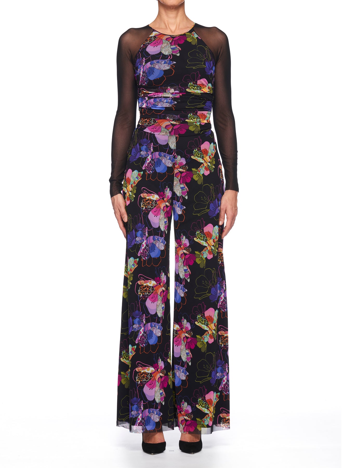 The individual is wearing the Fuzzi Woman Floral Wide Leg Jumpsuit from Fuzzi, which showcases a vibrant floral print on a black background. The top has sheer mesh fabric on the sleeves that complements the wide-leg silhouette as they stand against a plain white backdrop.