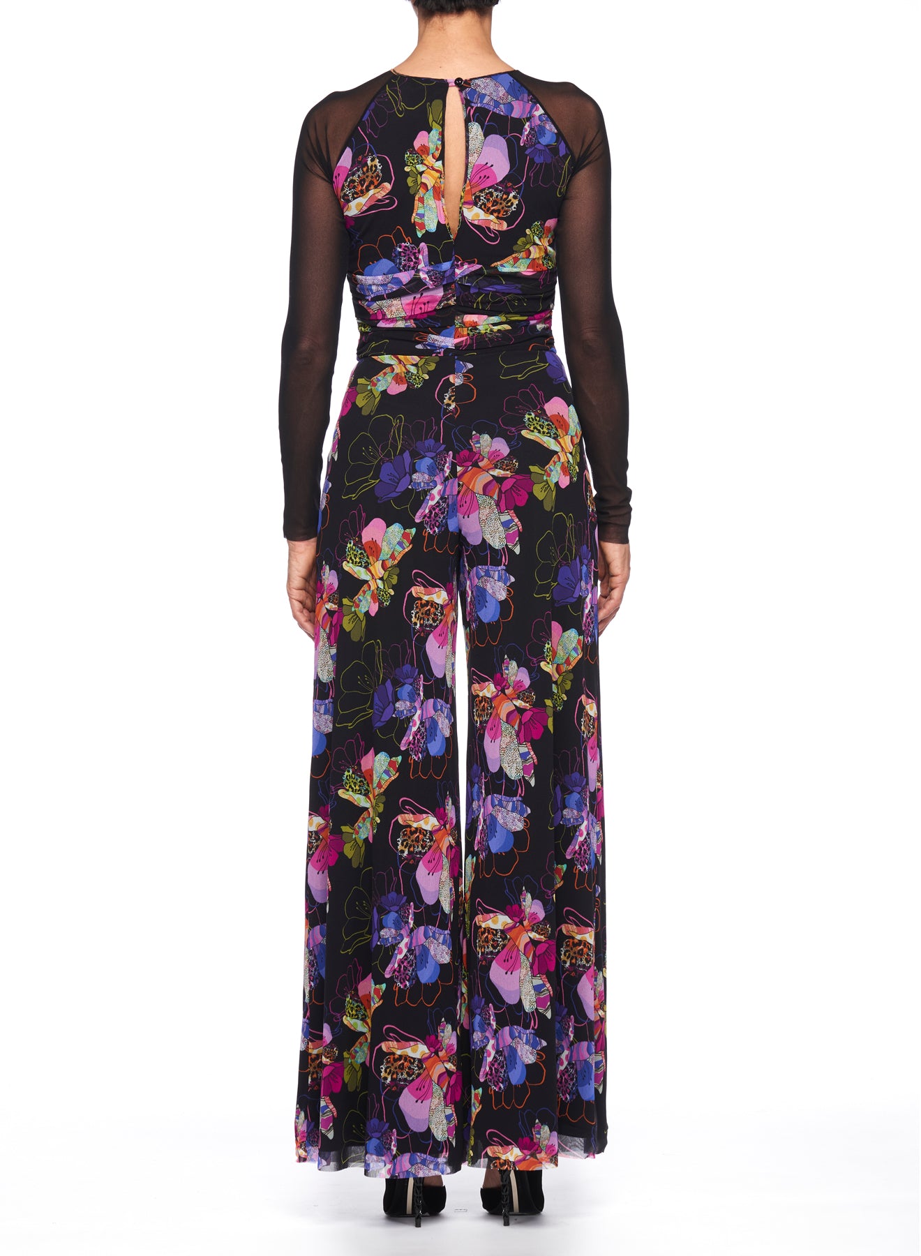 A rear view showcases a person wearing the Fuzzi Woman Floral Wide Leg Jumpsuit, adorned with a vibrant floral print. The jumpsuit features sheer mesh on the long sleeves and a keyhole back opening, set against a plain white background.