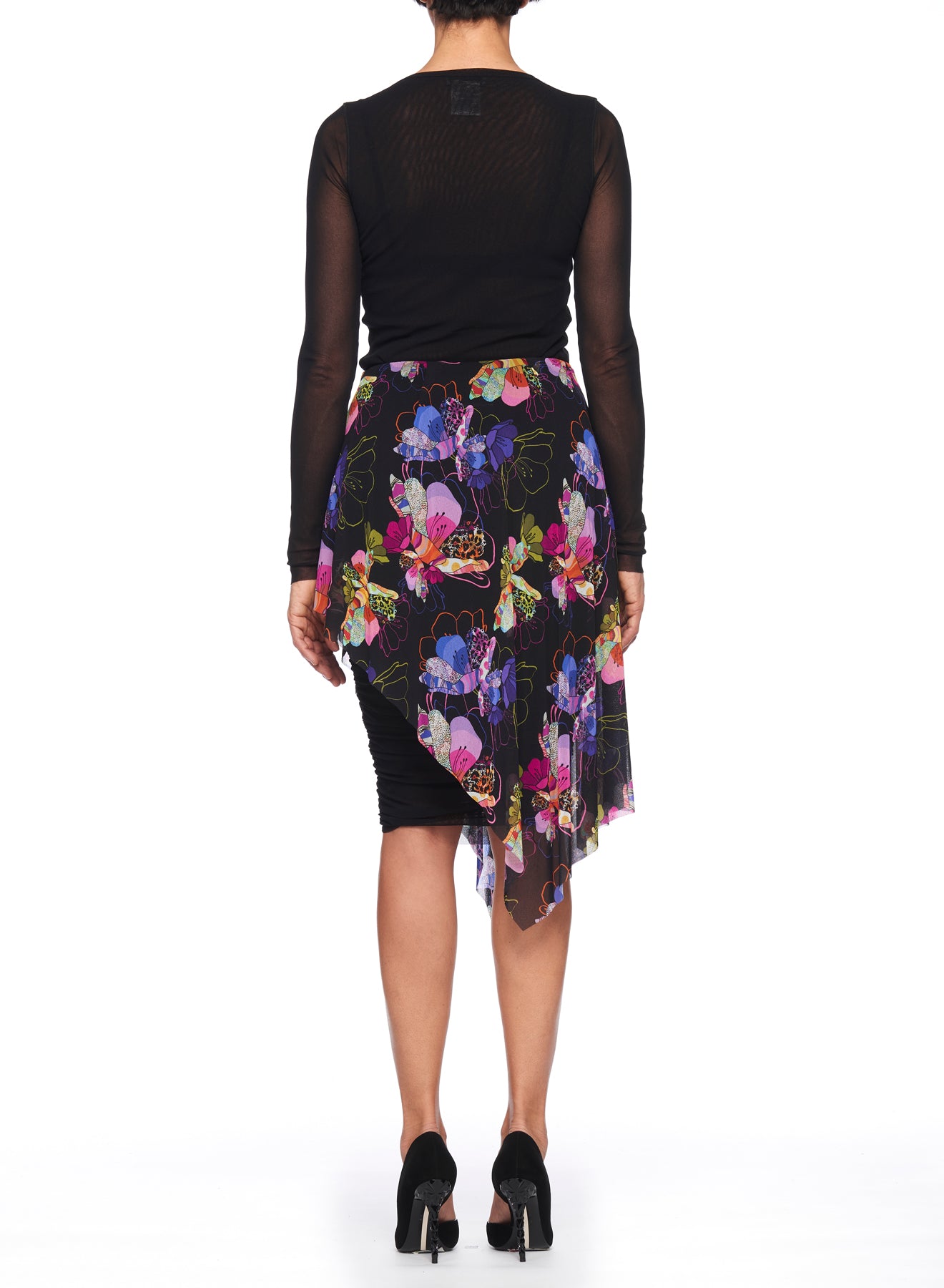 A person stands with their back to the camera, wearing a sheer black long-sleeve top and the Fuzzi Woman Asymmetrical Floral Mesh Skirt by Fuzzi, which showcases an asymmetrical hemline and vibrant colors. The look is completed with black high-heeled shoes against a plain white background.