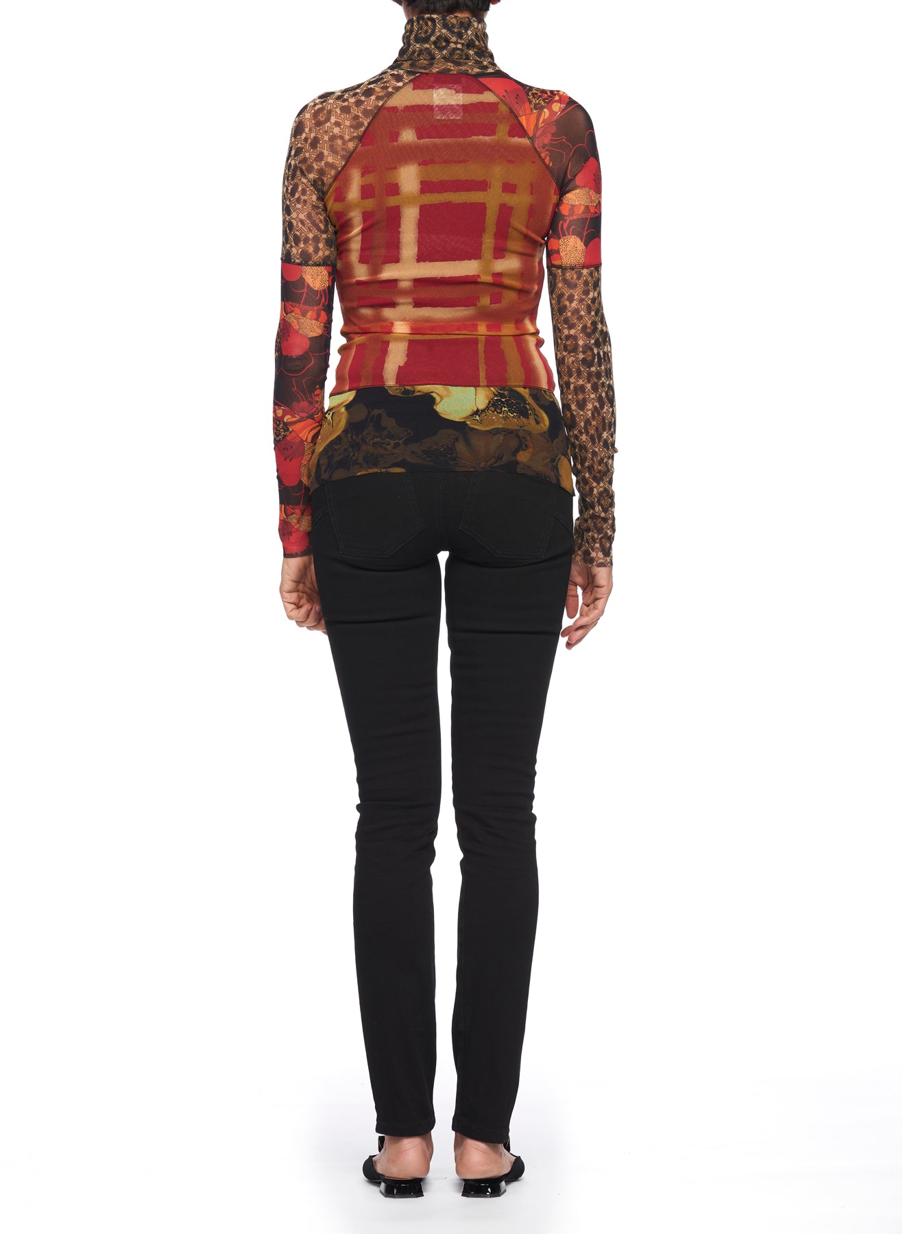 An individual wearing the Fuzzi Woman Bold Mixed-Print Mesh Turtleneck, which features a vibrant blend of floral, plaid, and leopard patterns reminiscent of a vintage style, is paired with black pants and heels. They are standing against a plain white background with their back to the camera.