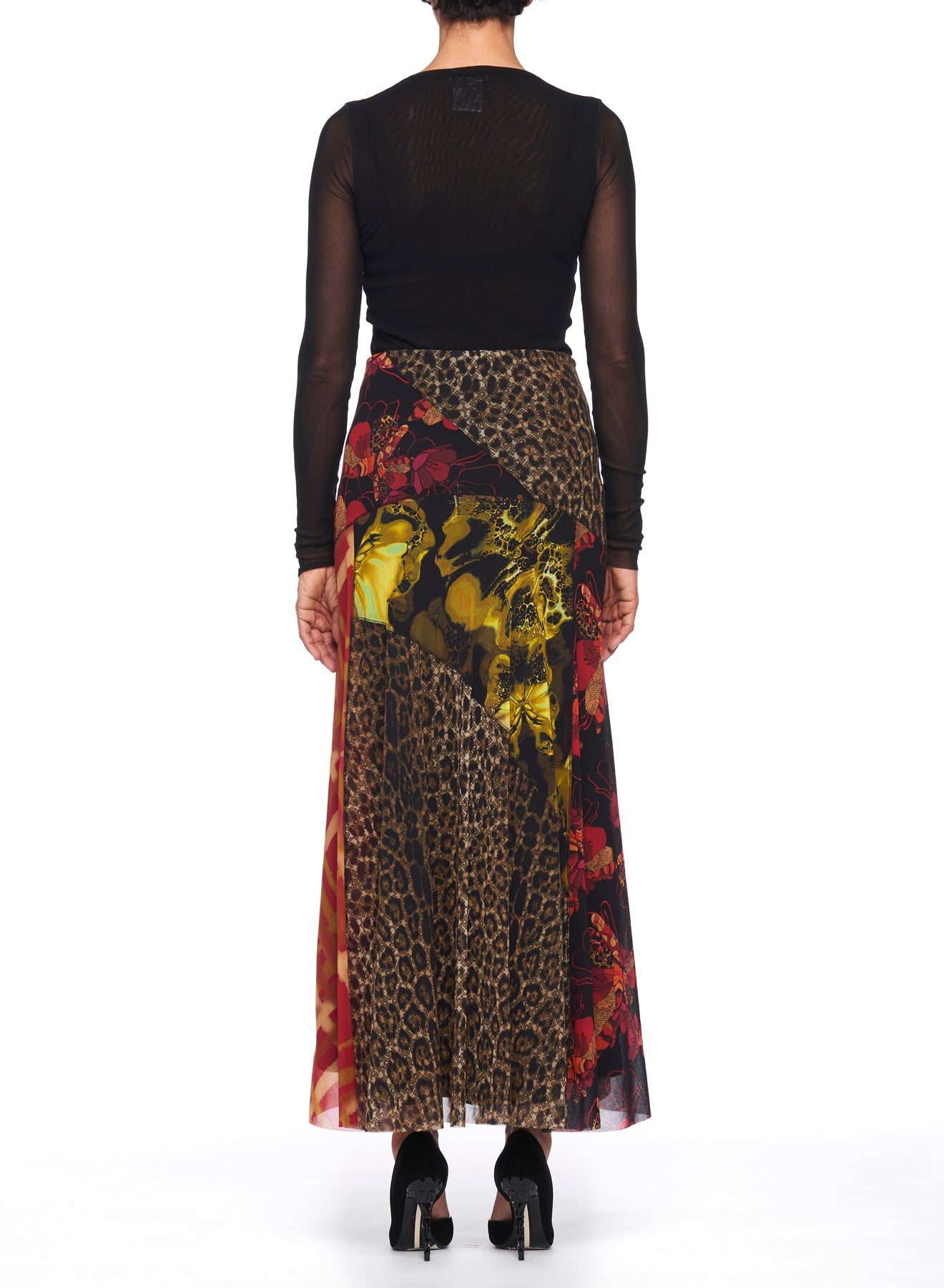 A person wearing a black sheer long-sleeve top and a vibrant, vintage piece stands facing away. The Fuzzi Woman Mixed-Print Mesh Long Skirt, adorned with animal and floral prints, features an asymmetrical hemline. They complete the look with black high heels.