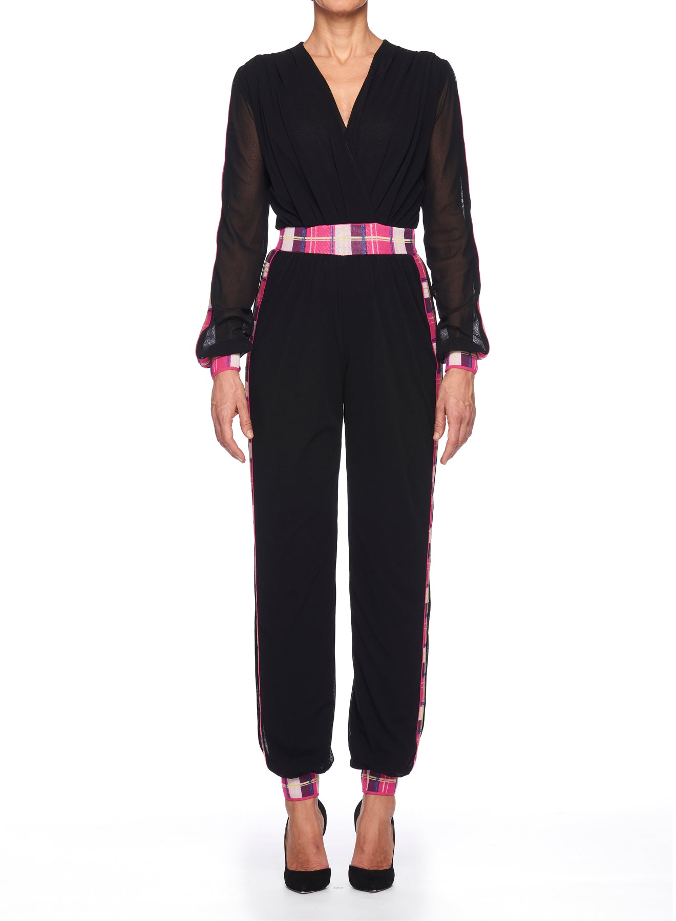 Wearing the Fuzzi Woman Black Mesh Jumpsuit with Colorful Details, which showcases sheer long sleeves and plaid accents in pink, red, and white at the waist, sides, and cuffs, a person completes their ensemble with black high-heeled shoes against a pristine white background.