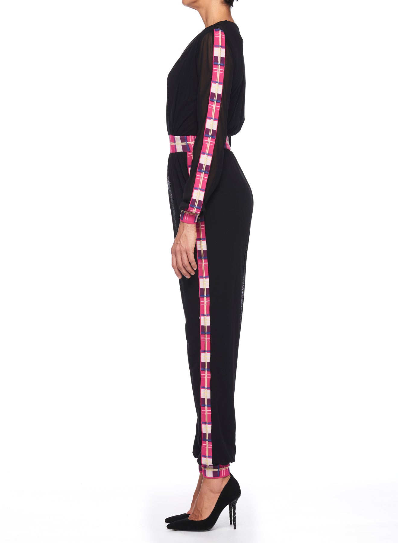 A person in side profile wears the Fuzzi Woman Black Mesh Jumpsuit with Colorful Details, featuring sleek black mesh with vibrant pink plaid accents along the sides and sleeves, elegantly paired with black high-heeled shoes. The crisp white background accentuates the outfit's striking details.