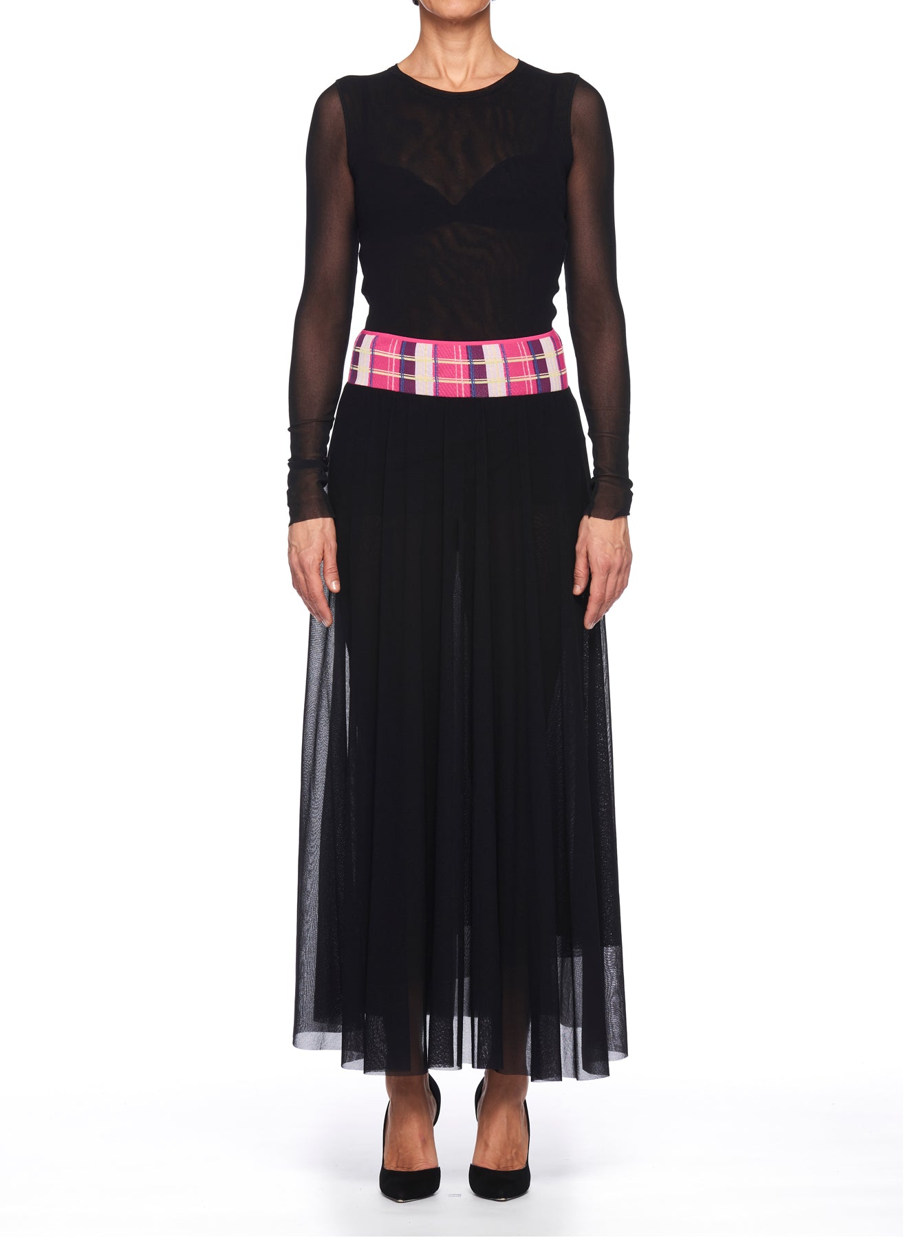 A person stands against a white background wearing the Fuzzi Women's black mesh skirt with sheer long sleeves and a midi length. The ensemble is complemented by a colorful plaid pink and purple belt that adds vintage charm. They complete the look with black high heels.