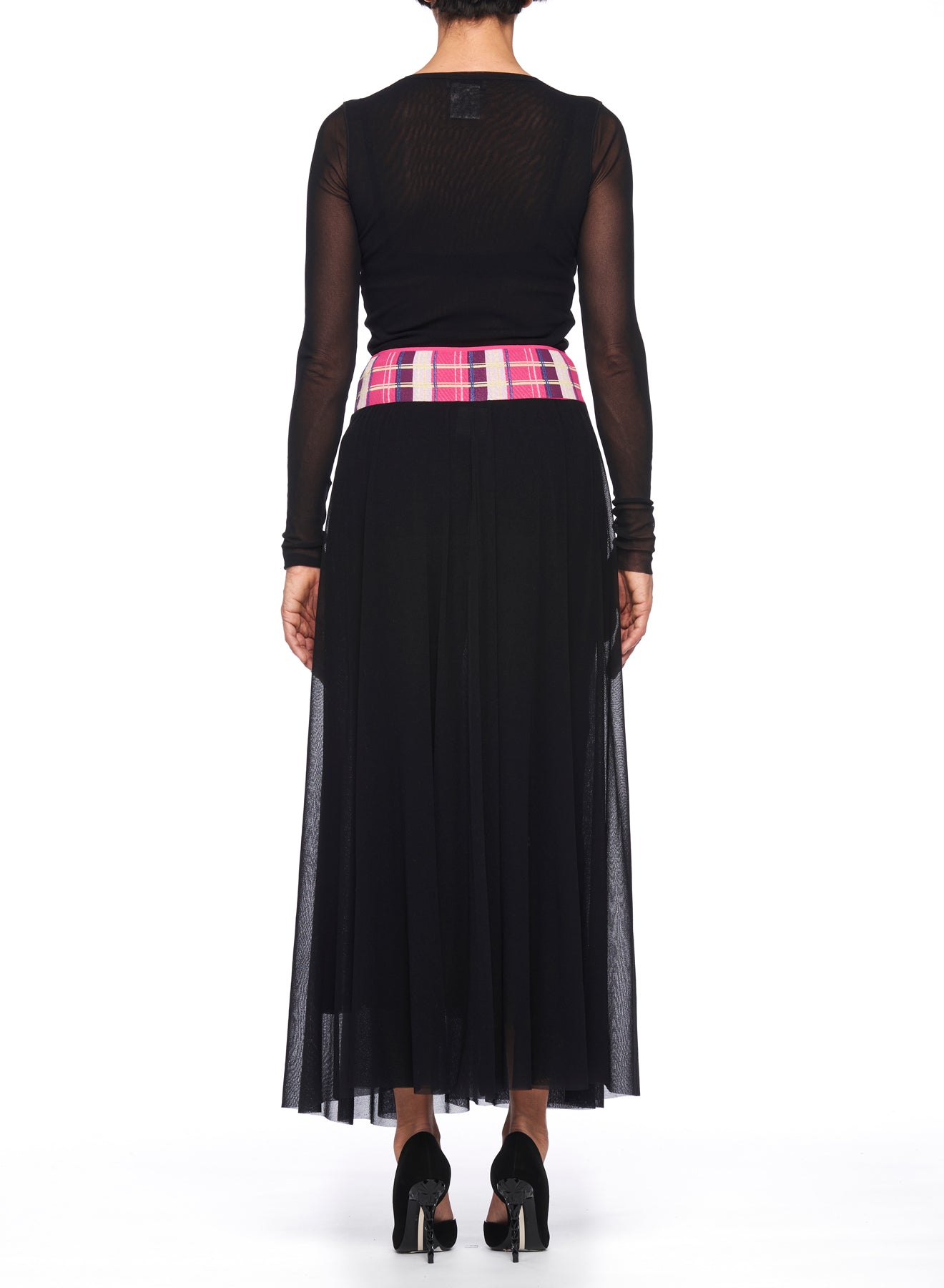 A person stands facing away, radiating vintage charm in a Fuzzi black mesh midi skirt with a colorful contrasting belt and a sheer long-sleeve black top. Black high-heeled shoes complete the look against a white background.