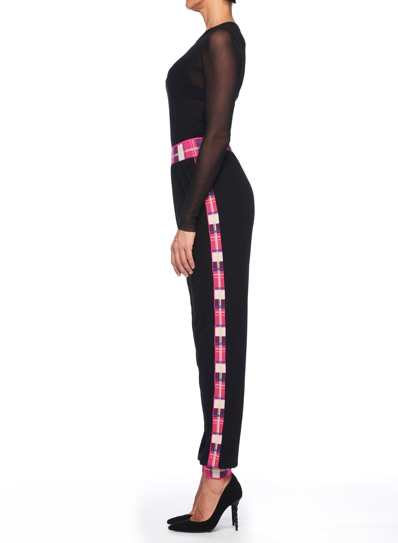 The image displays a side profile of an individual wearing Fuzzi Woman Black Pants with Colorful Side Stripes, highlighted by a pink plaid design. The ensemble is completed with a black long-sleeve top and chic black heels, all presented against a clean white backdrop.