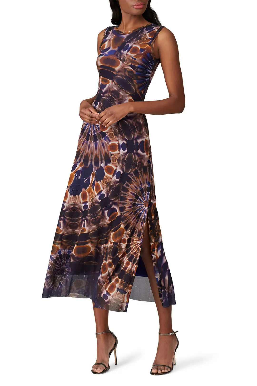 A woman is wearing the Fuzzi Woman Tie-Dye Mesh Midi Dress, which has a sleeveless design and an ankle-length silhouette featuring a bold and intricate abstract pattern in brown and navy. The dress includes a side slit, which she effortlessly pairs with strappy high-heeled sandals.