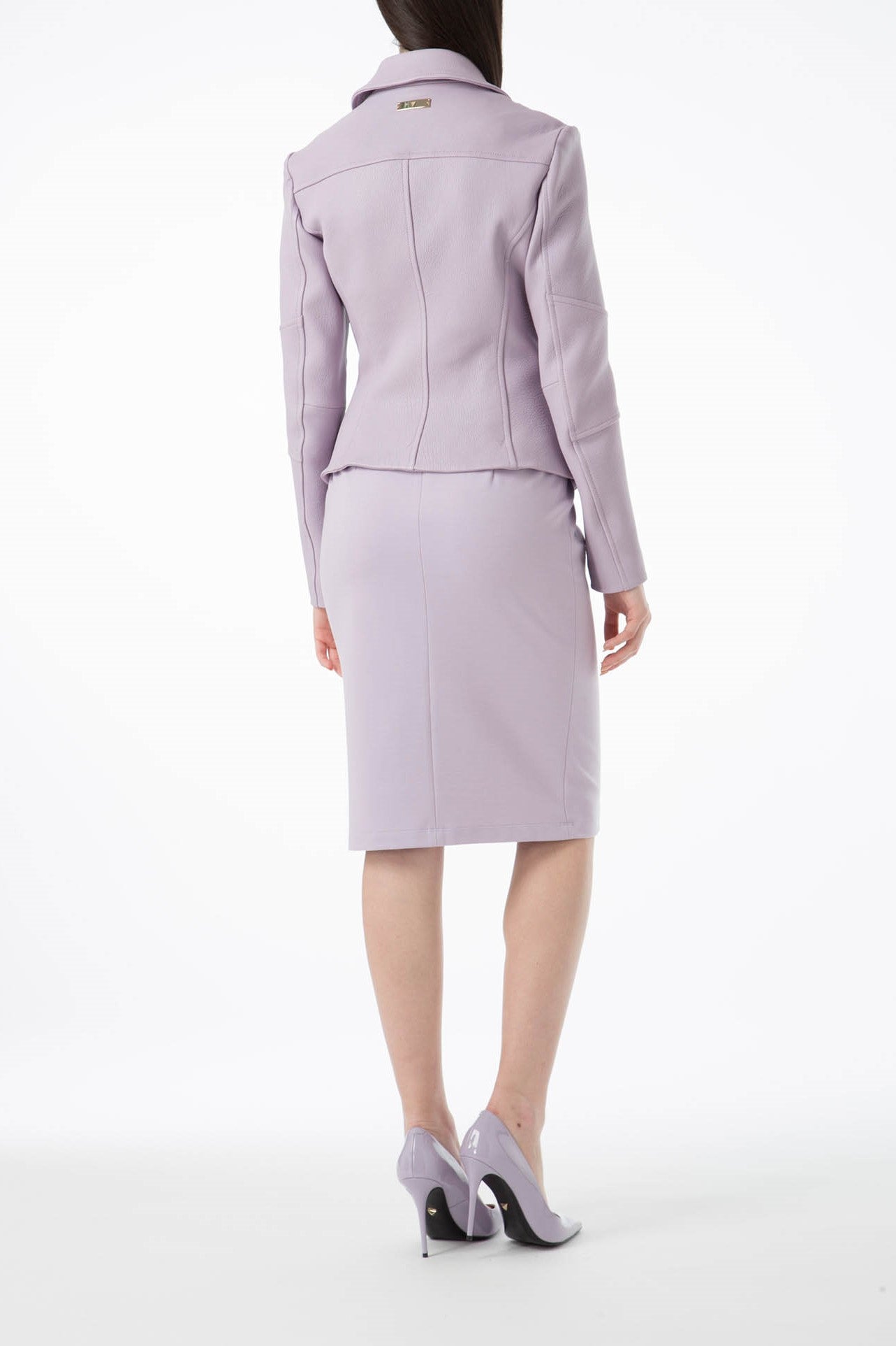 A person stands with their back turned, dressed in the Antonio Croce Ginette Jacket featuring gold hardware, paired with a light lavender skirt suit and matching high-heeled shoes. The elegant jacket from Antonio Croce completes a classic look against the plain white backdrop, highlighting the outfit's color and design.