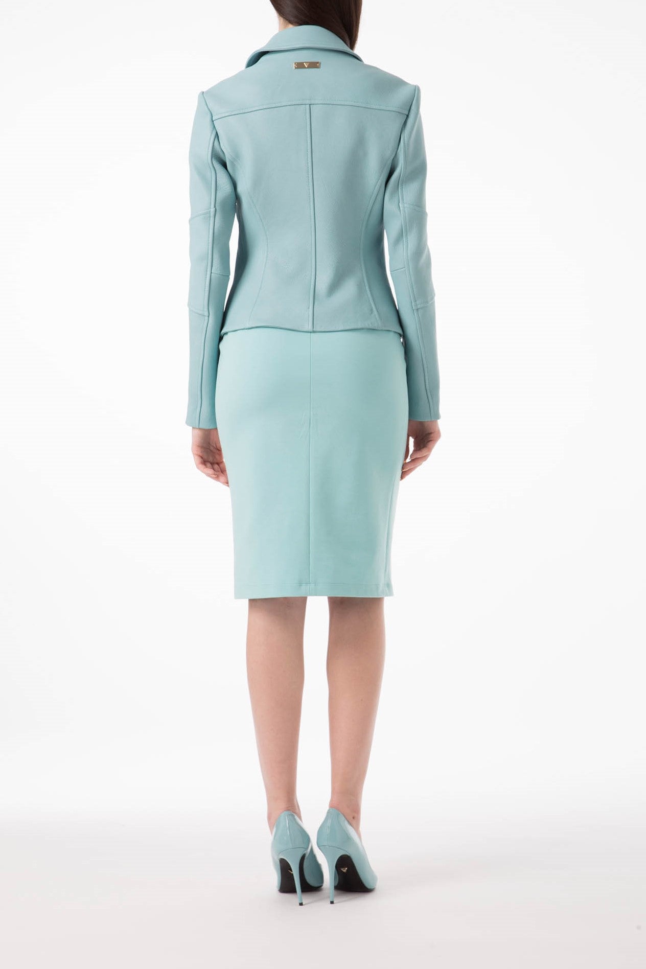 A woman stands with her back to the camera, wearing the Antonio Croce Ginette Jacket with Gold Hardware, paired with a matching knee-length skirt and high heels. The plain white backdrop highlights her poised style.