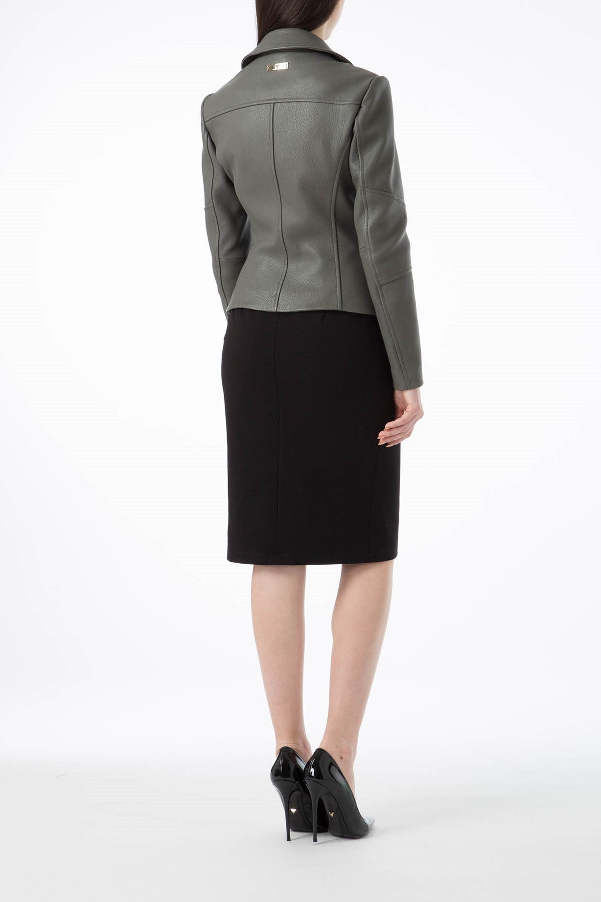 An individual wearing an Antonio Croce Ginette Jacket, featuring elegant dark gray leather with subtle gold hardware, pairs it with a black pencil skirt. Complemented by black high-heeled shoes, this vintage-inspired ensemble is set against a plain white background.
