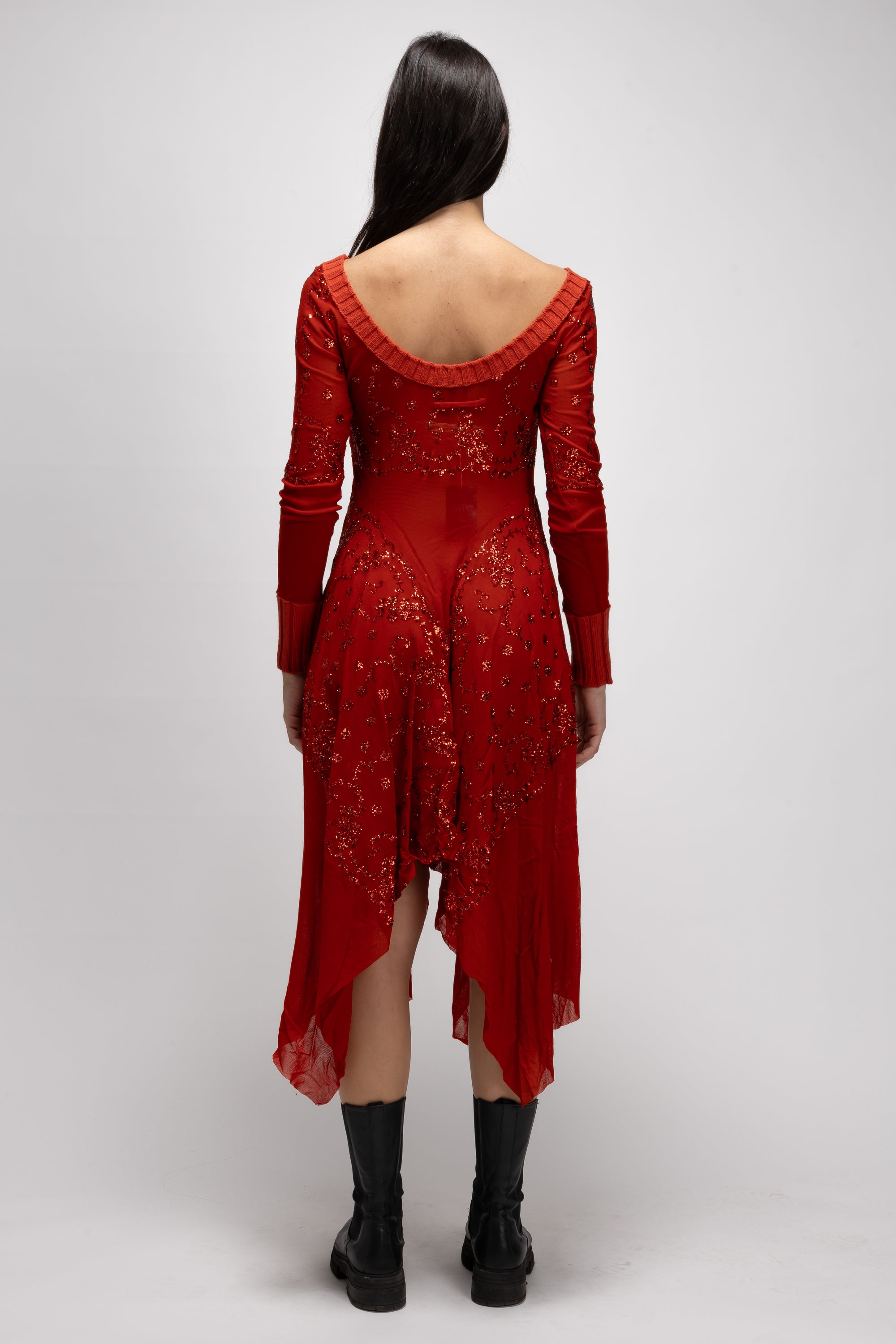 A person with long dark hair is wearing a Vintage Woman Red Dress by Jean Paul Gaultier from the Spring/Summer 1995 collection. The red dress, adorned with intricate patterns and a low back, highlights its high-low hem. They stand against a plain gray background and complete the look with black ankle boots.